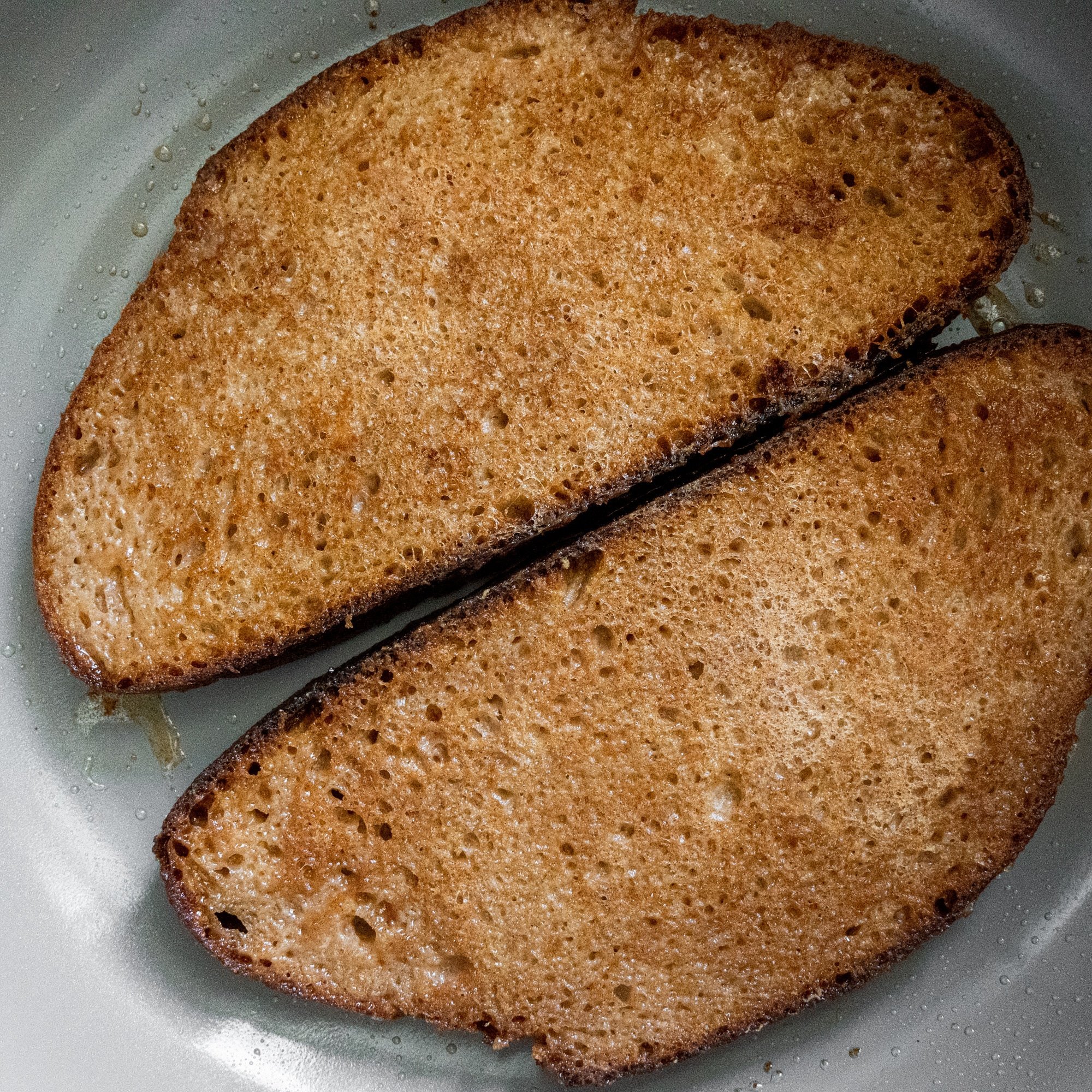 A close up of two pieces of french toast golden on top laid side by side in a grey frying pan.
