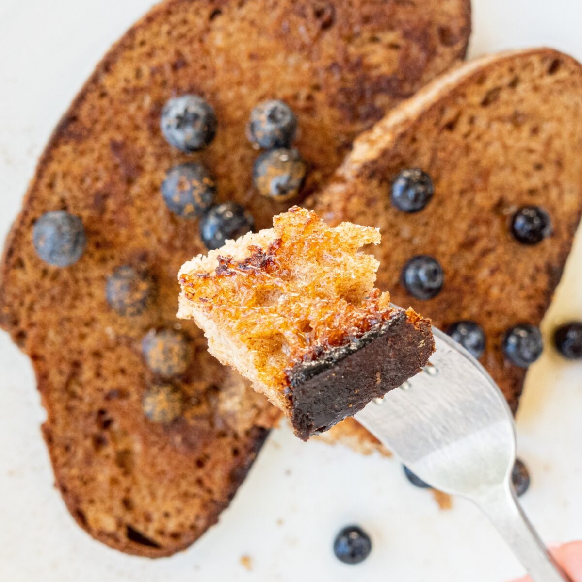 Two pieces of vegan French toast with maple syrup and blueberries on top. A fork is held in the center of the photo with a bite of French toast on the end.