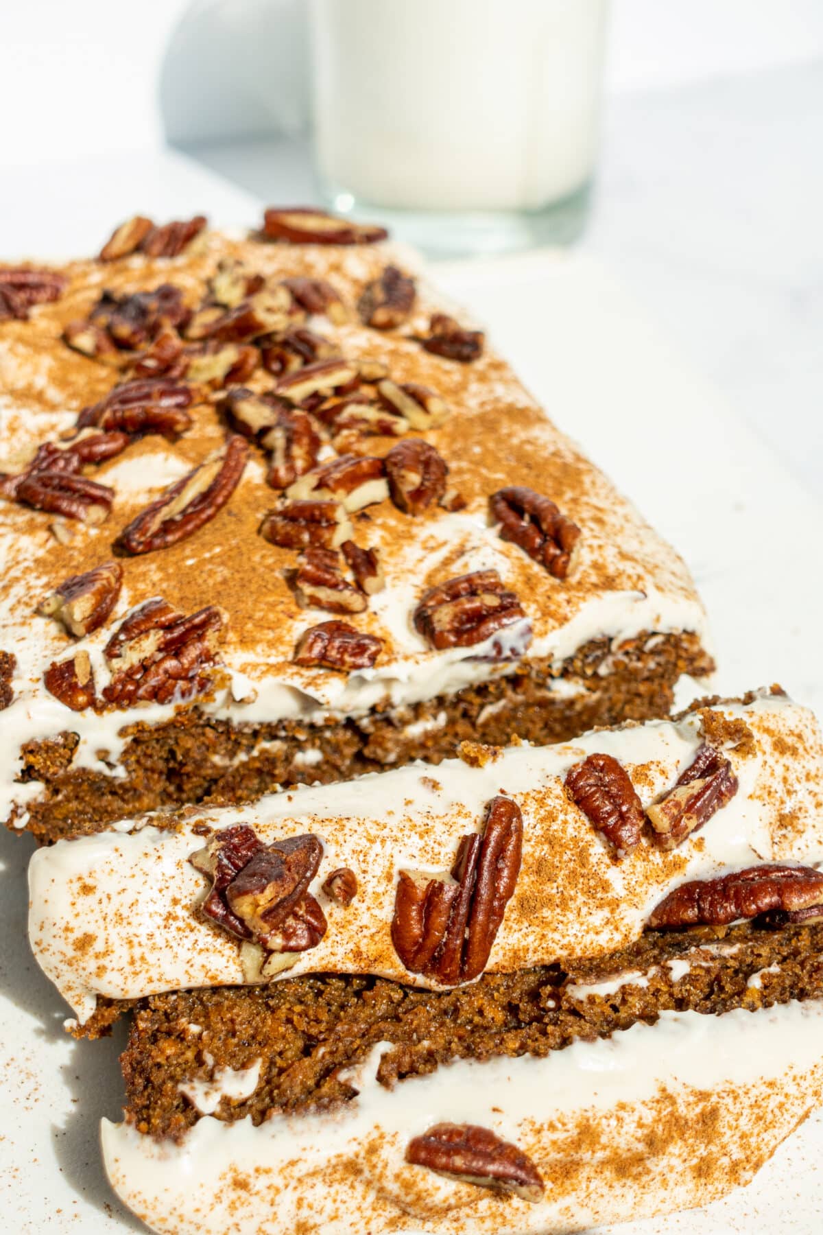 A loaf of vegan gluten free carrot cake with two slices cut off at the end. In the background is a glass of milk.