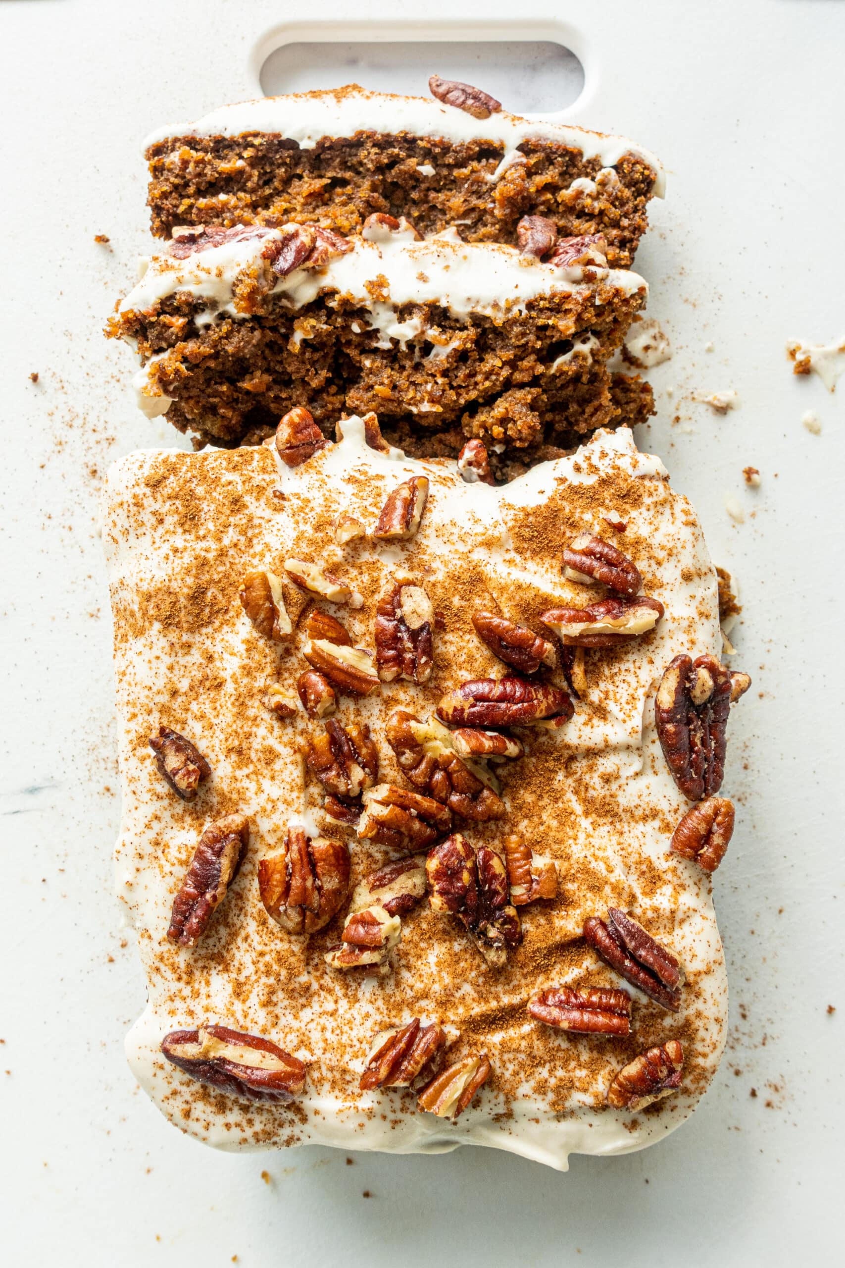 A loaf of vegan gluten free carrot cake topped with vegan cream cheese frosting, cinnamon, and chopped pecans on a white cutting board with the first two pieces cut.