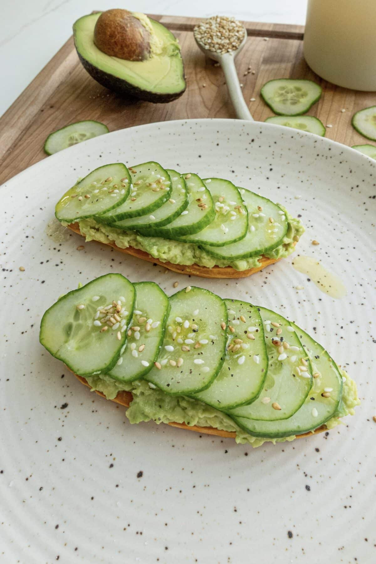 Two pieces of Sweet Potato Avocado Toast topped with cucumber and sesame seeds on a white plate. Behind the plate is a brown wooden cutting board with half an avocado on it, a small spoon filled with sesame seeds, and more cucumber slices.