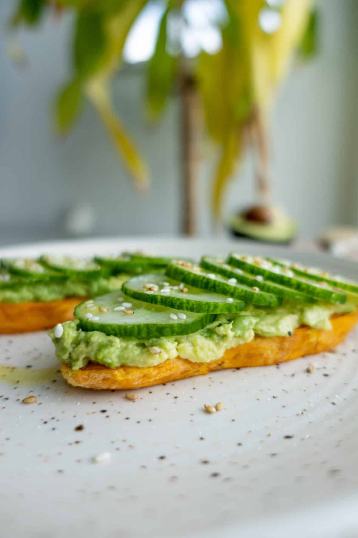 A close up of two pieces of sweet potato toast topped with mashed avocado and sliced cucumber, garnished with sesame seeds.