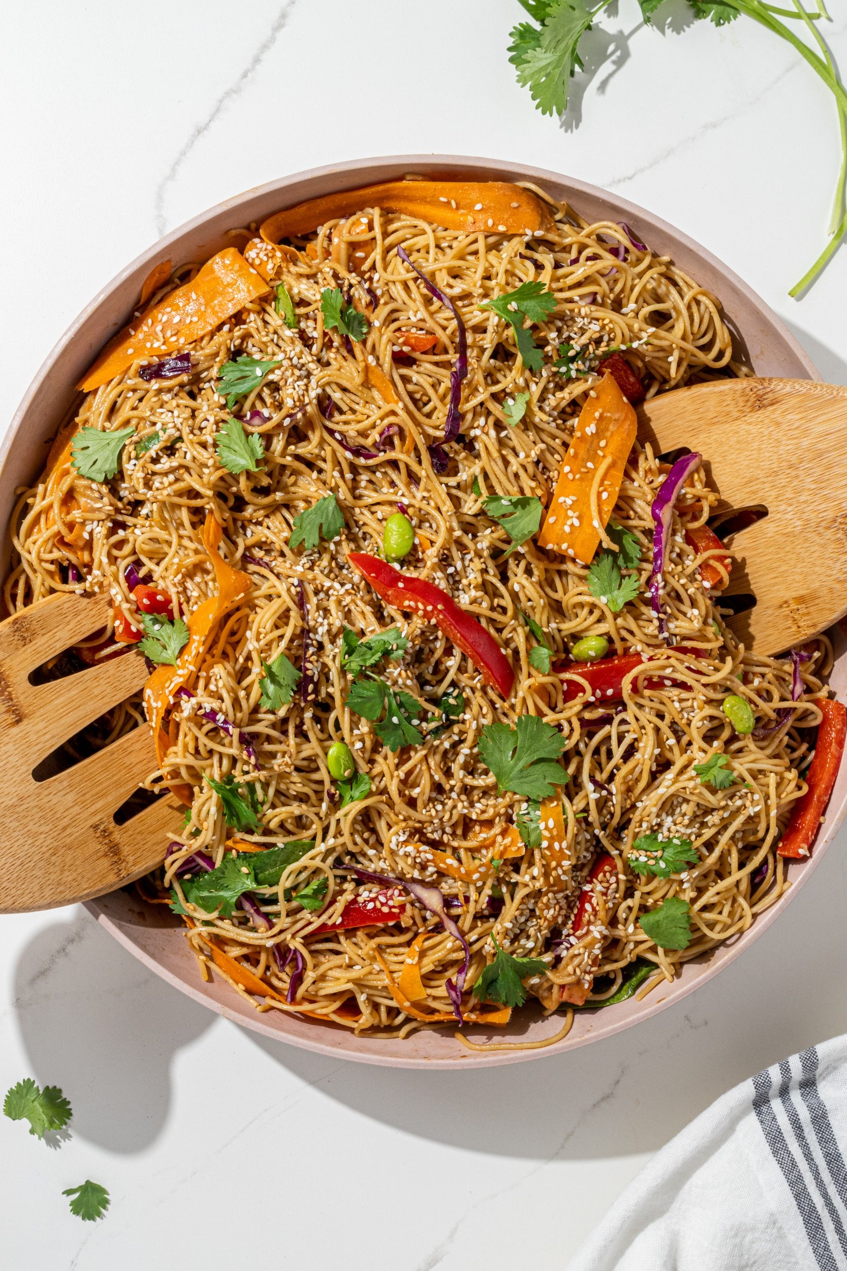 A white bowl filled with vegan peanut noodles and two brown wooden tongs sitting on the edges of the bowl. Surrounding the bowl is cilantro leaves and a white towel for garnish.