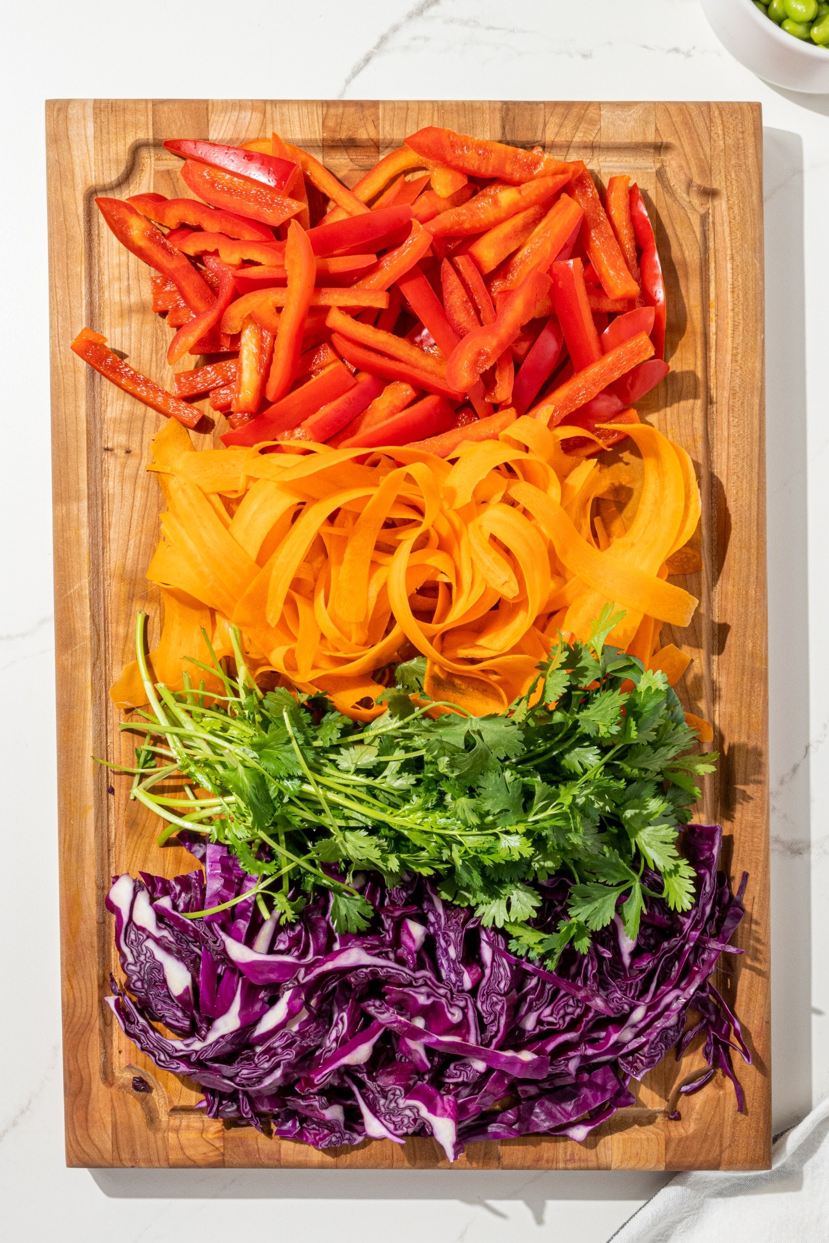 A brown wood cutting board filled with red pepper, carrot, cilantro, and purple cabbage all chopped.