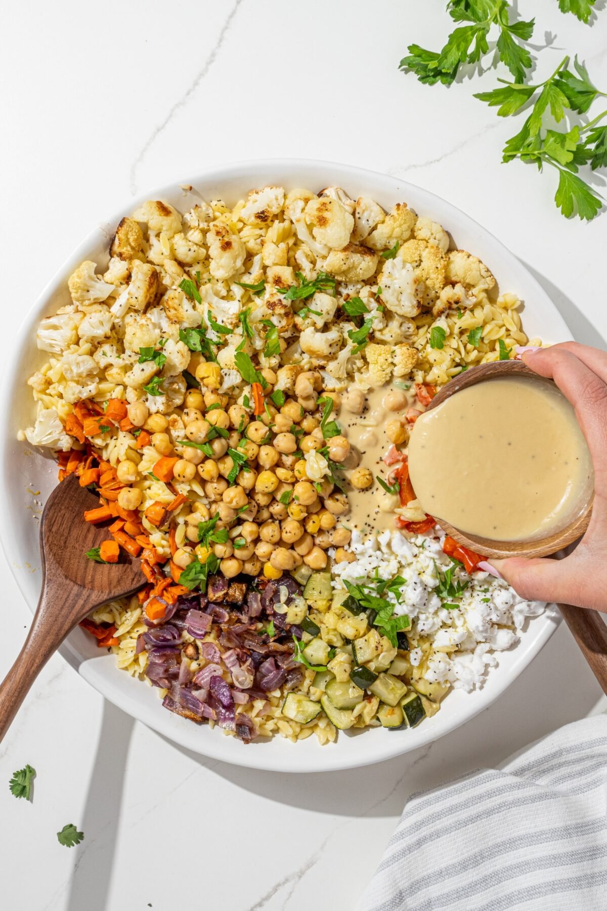 A large white bowl of roasted vegetable orzo salad with a pair of tongs in the bowl. A white hand is coming in from the right of the image pouring hummus salad dressing from a small bowl.