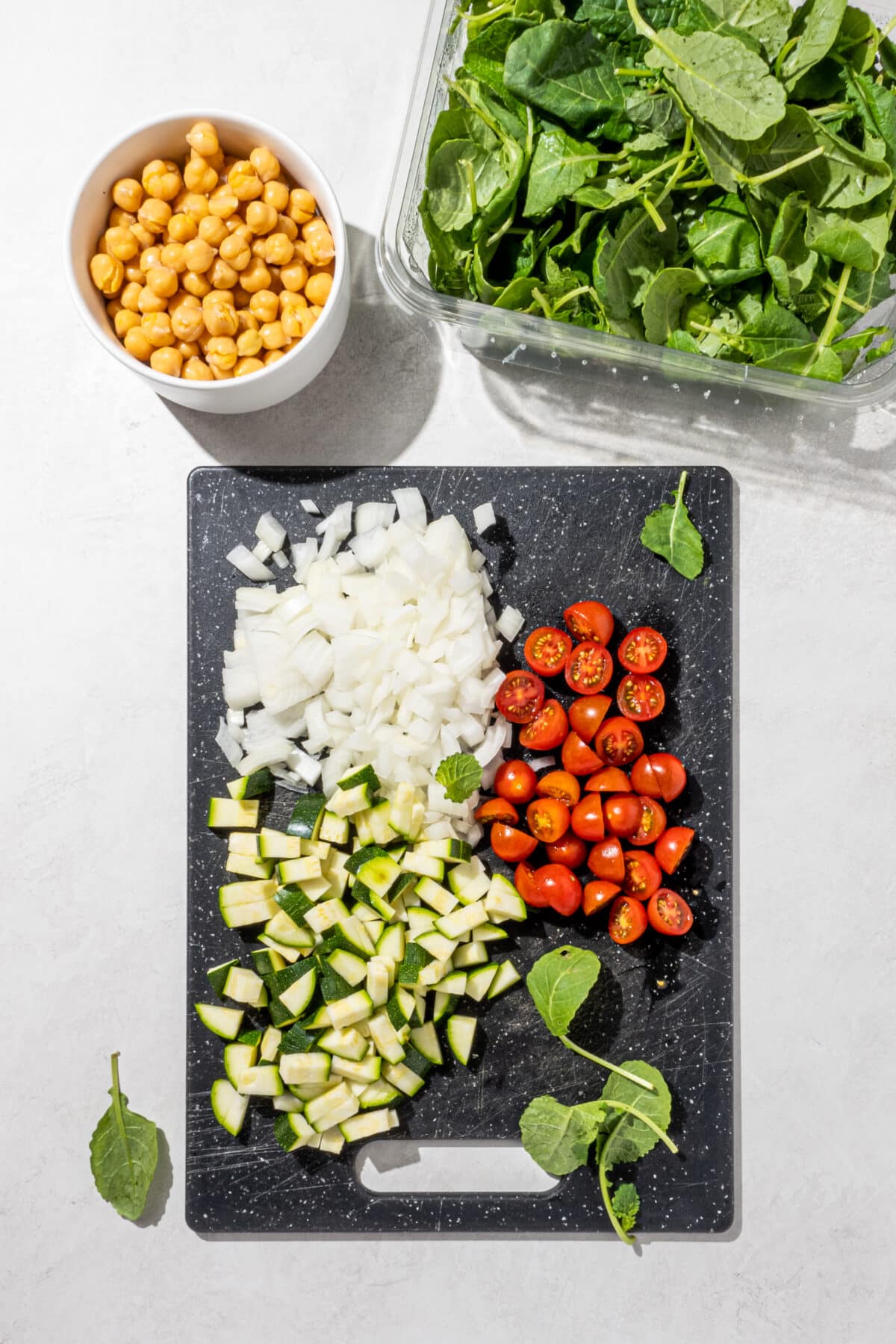 A black cutting board with chopped onions, zuchinni, and tomatoes on it along with a clear box of spinach and a white bowl of chickpeas.