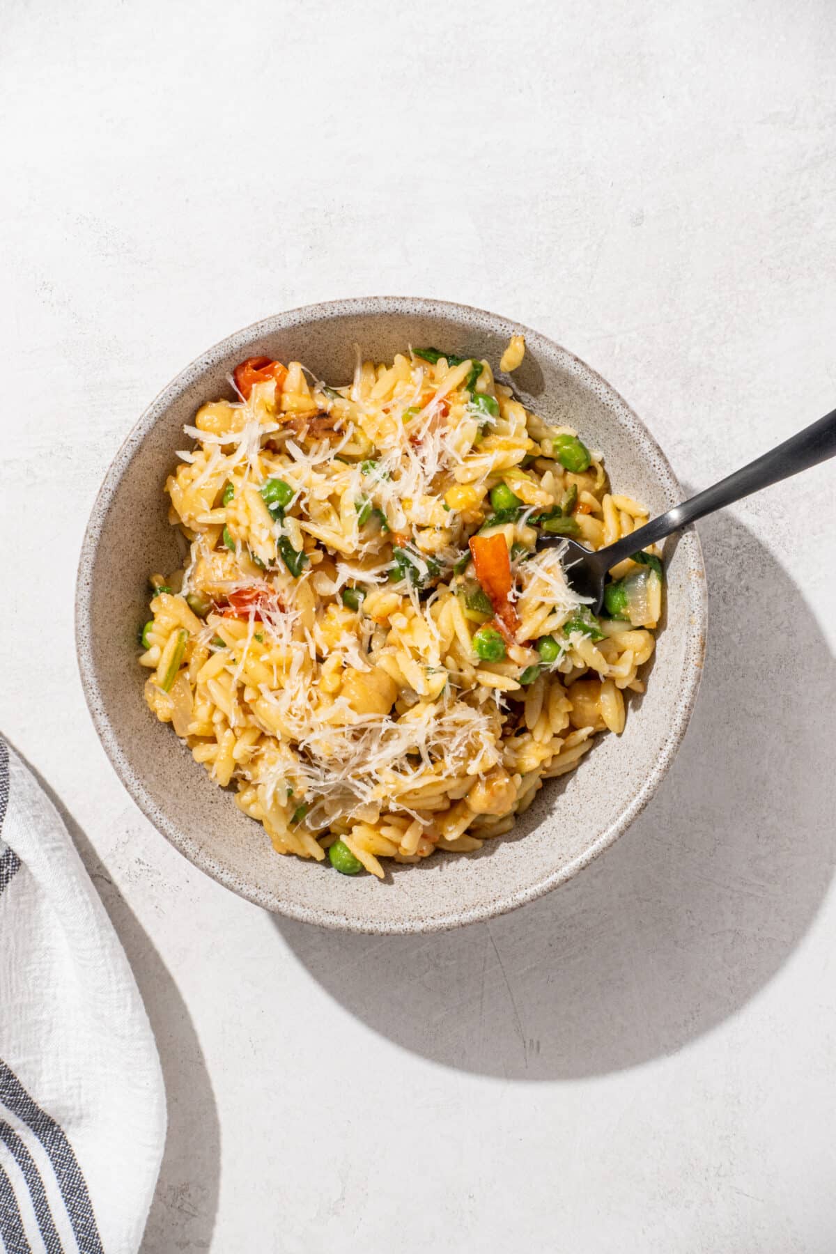 A single bowl of one pot healthy orzo garnished with parmesan cheese and a black fork sticking out of the bowl. To the left of the bowl is a black and white striped towel.