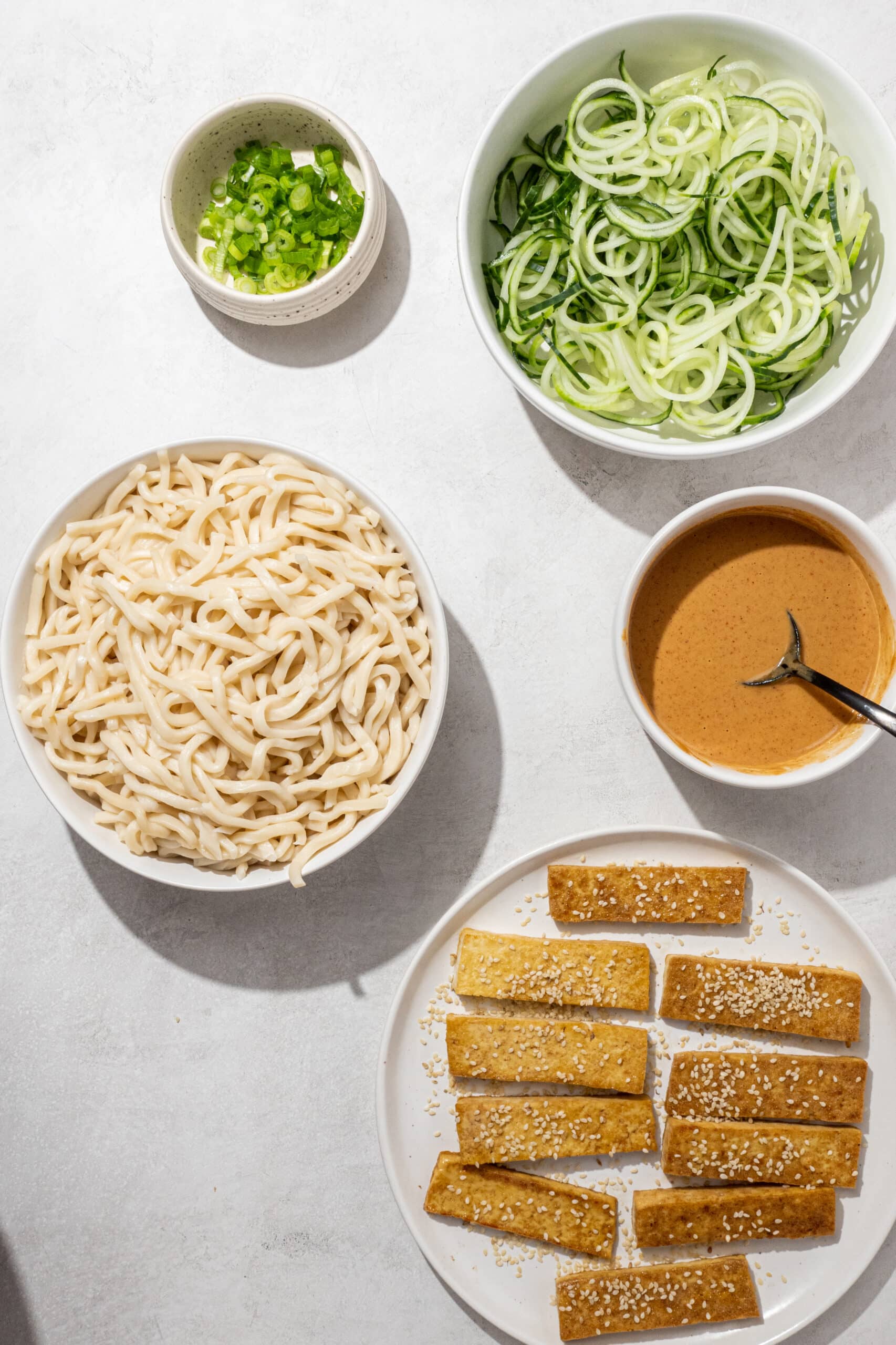 Ingredients needed to make peanut udon noodles laid out on a marble counter and in bowls including: scallions, cucumber, peanut sauce, udon noodles, and tofu.