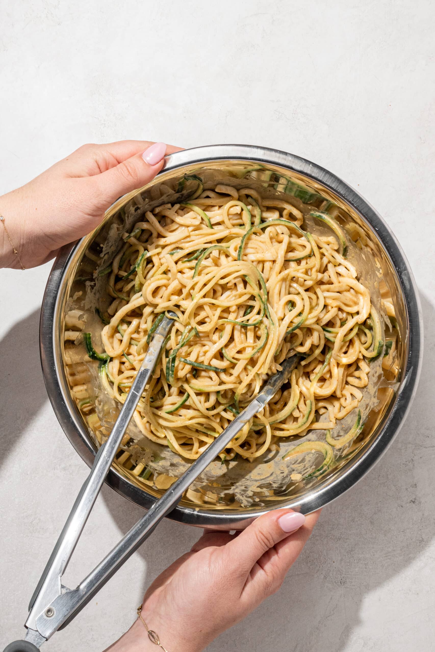 Two white hands holding the sides of a silver large bowl filled with peanut udon noodles and a pair of silver tongs.