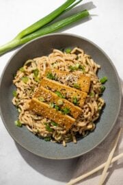 Easy Peanut Udon Noodles topped with tofu, green onion, and sesame seeds on a gray plate. To the right is a tan linen napkin and a set of chopsticks. On the left of the bowl is a stalk of green onion.