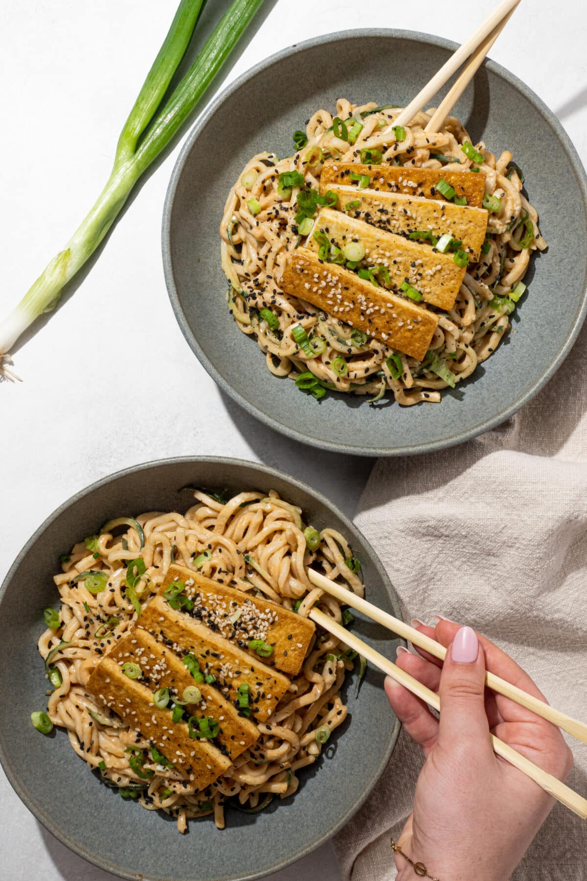 Two bowls of easy peanut udon noodles with tofu on top with a pair of chopsticks in each. In the bottom bowl, a white hand is holding the chopsticks in the noodles. To the left of the bowls is a green onion stalk and to the right is a tan linen napkin.