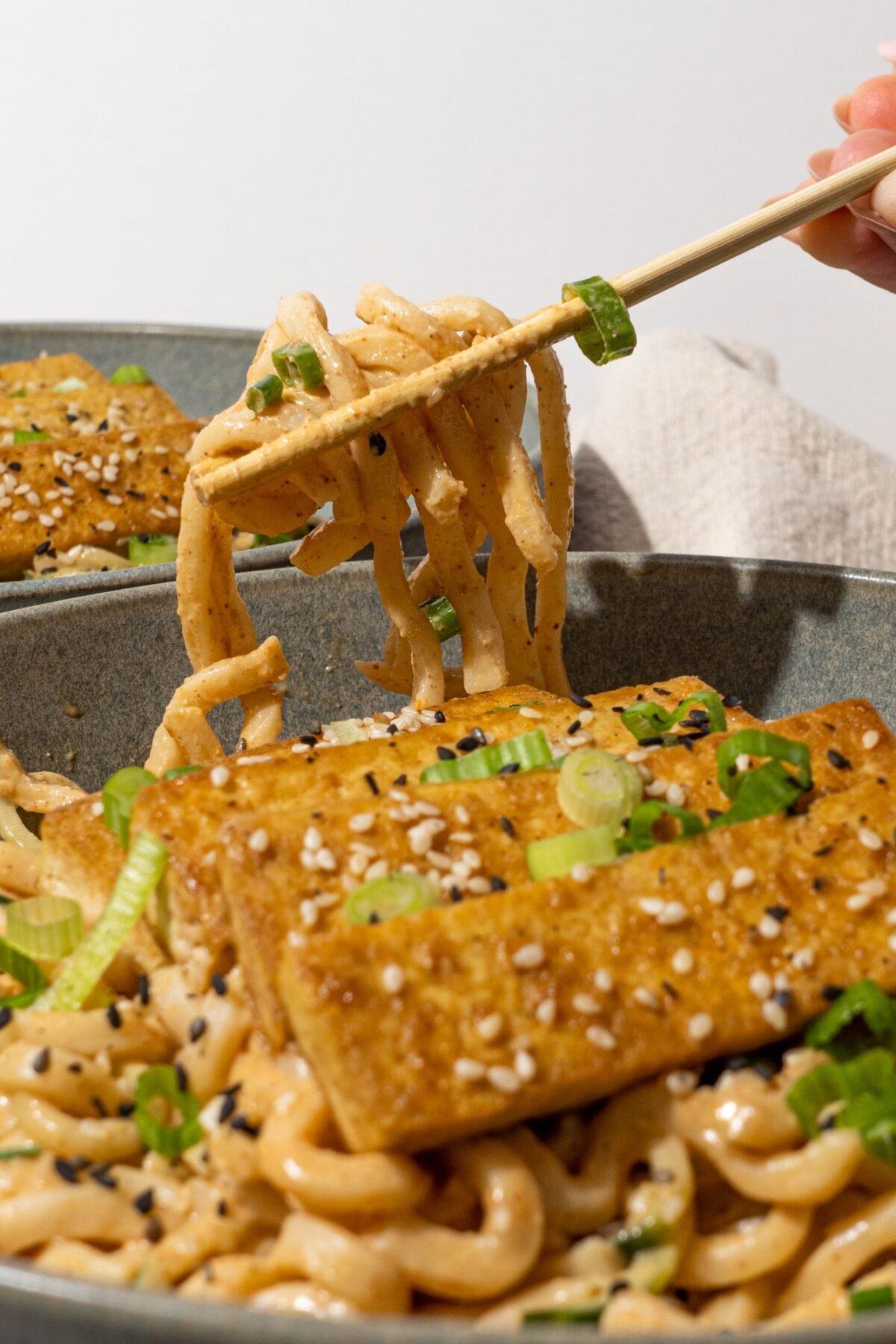 Close up of the easy peanut udon noodles in a gray bowl with a pair of chopsticks in the center holding a bite of noodles.