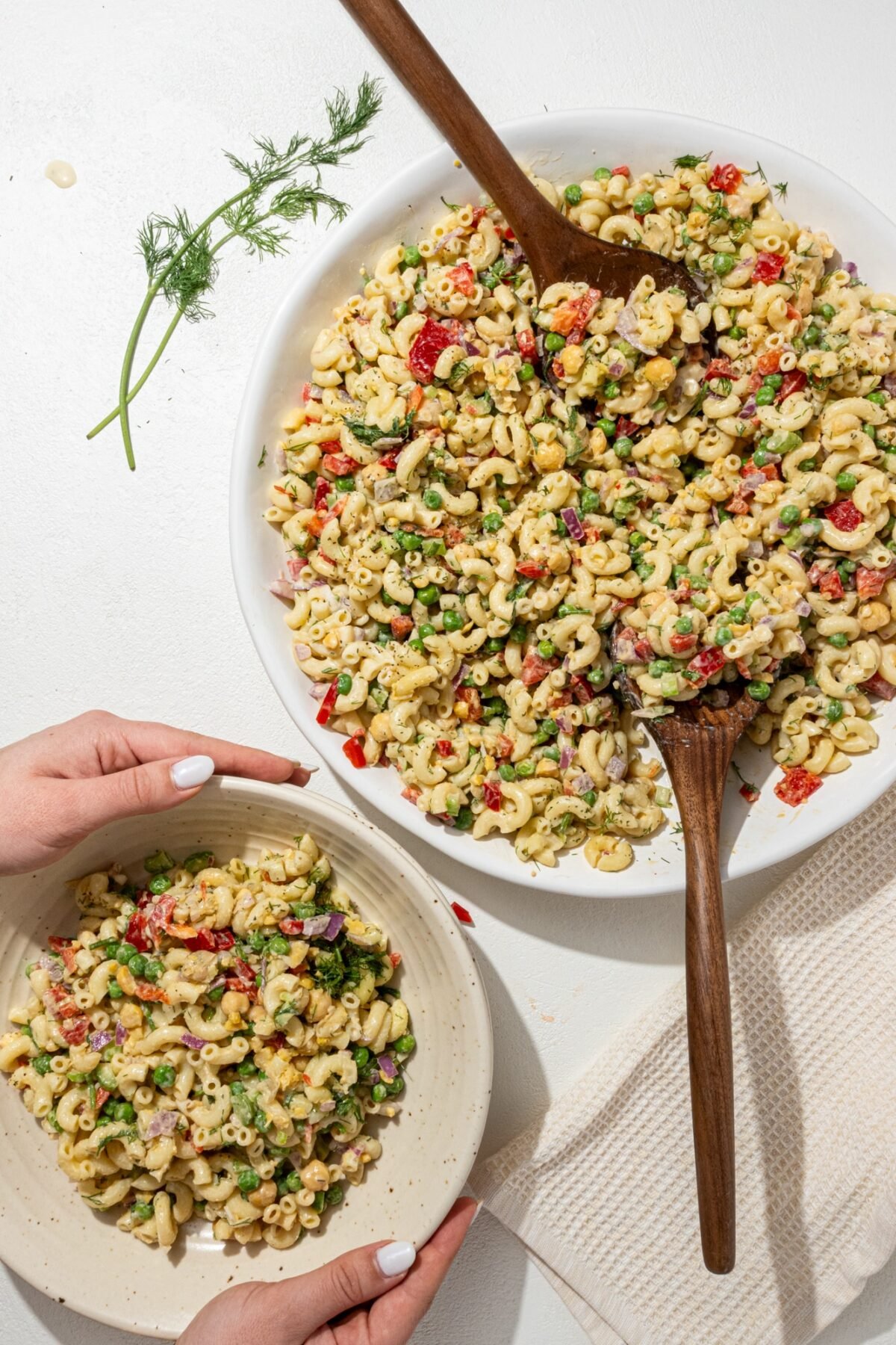 A large white serving bowl of tuna pasta salad with two wooden salad tongs in it. To the left is a small single serving bowl of tuna pasta salad and a sprig of fresh dill.
