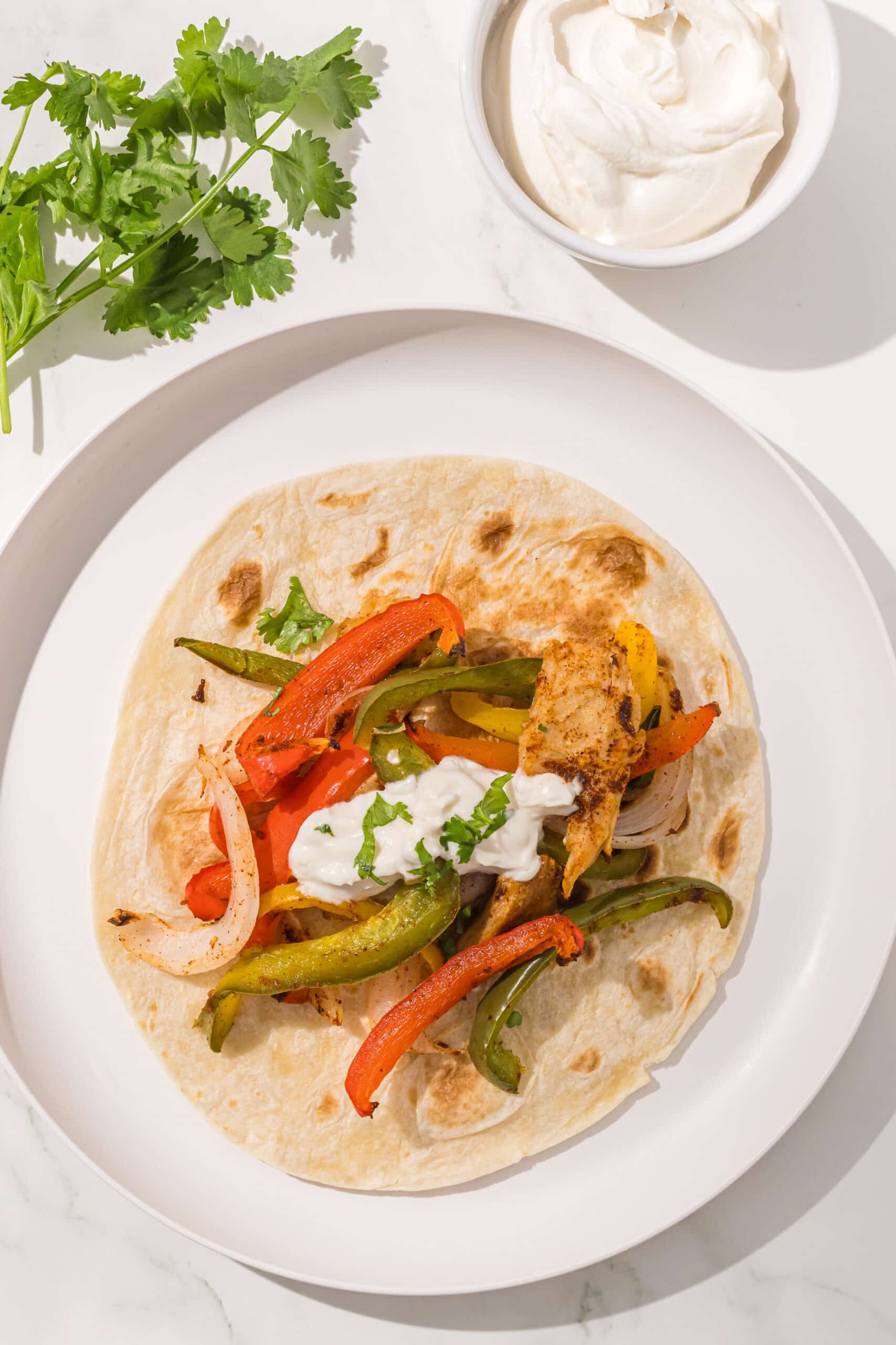 A single fajita on a white plate which is a tortilla with bell peppers, onions, chicken, and sour cream on it. Beside the plate is a pile of fresh cilantro and a small bowl of sour cream.