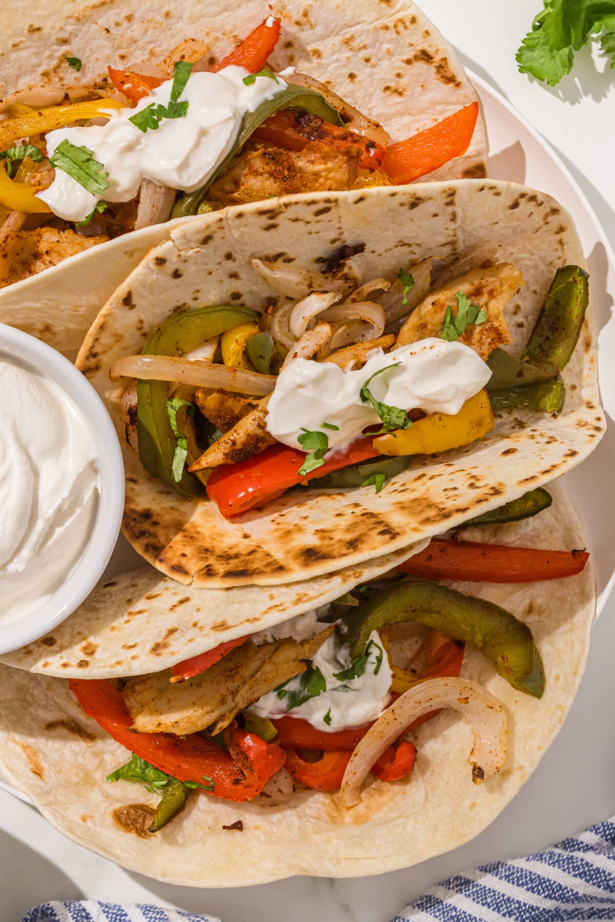 Three fajita tacos on a plate topped with sour cream and cilantro and a small bowl of sour cream.