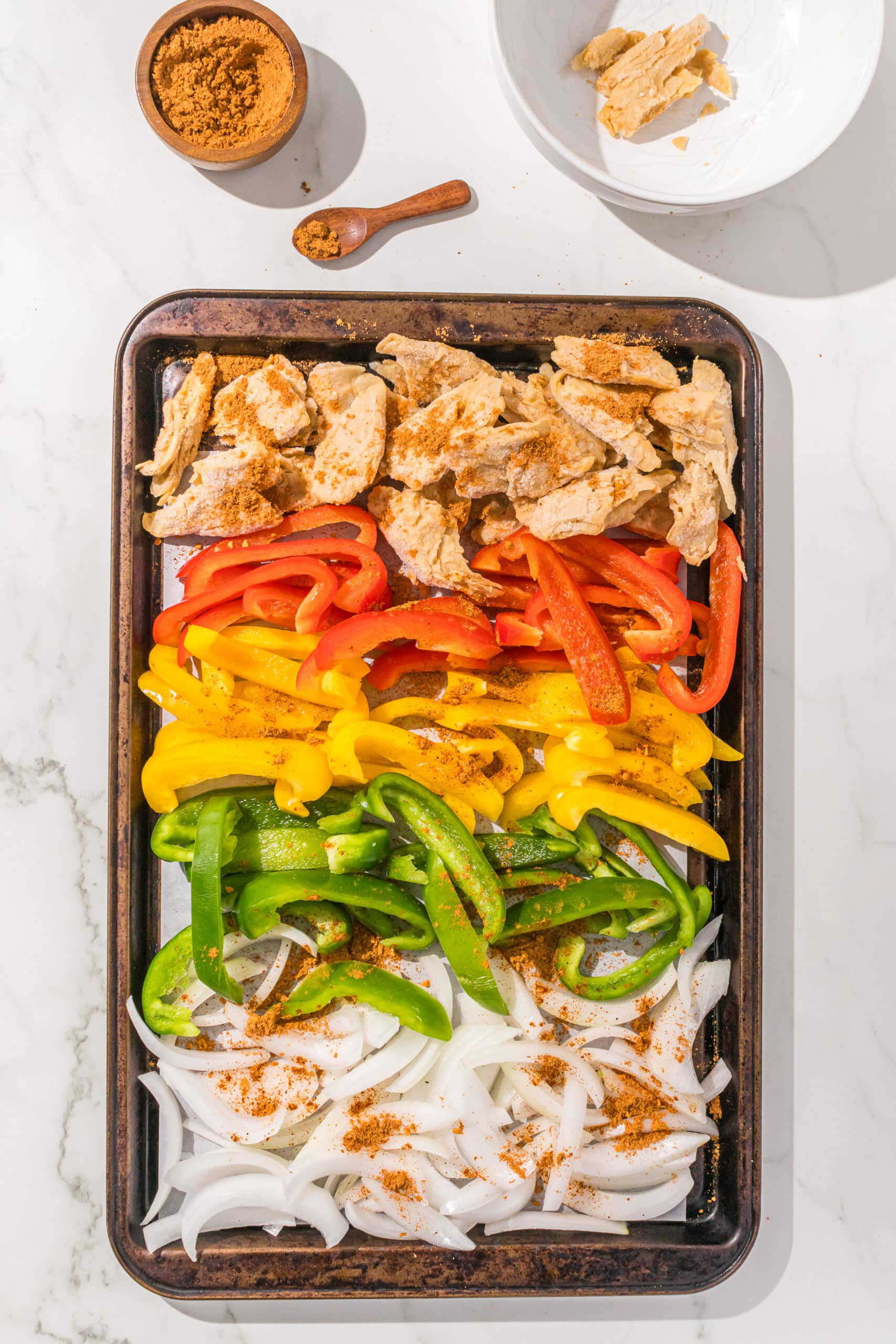 A sheet pan filled with sliced onions, bell peppers, and vegan chicken pieces and a small wooden bowl of taco seasoning.