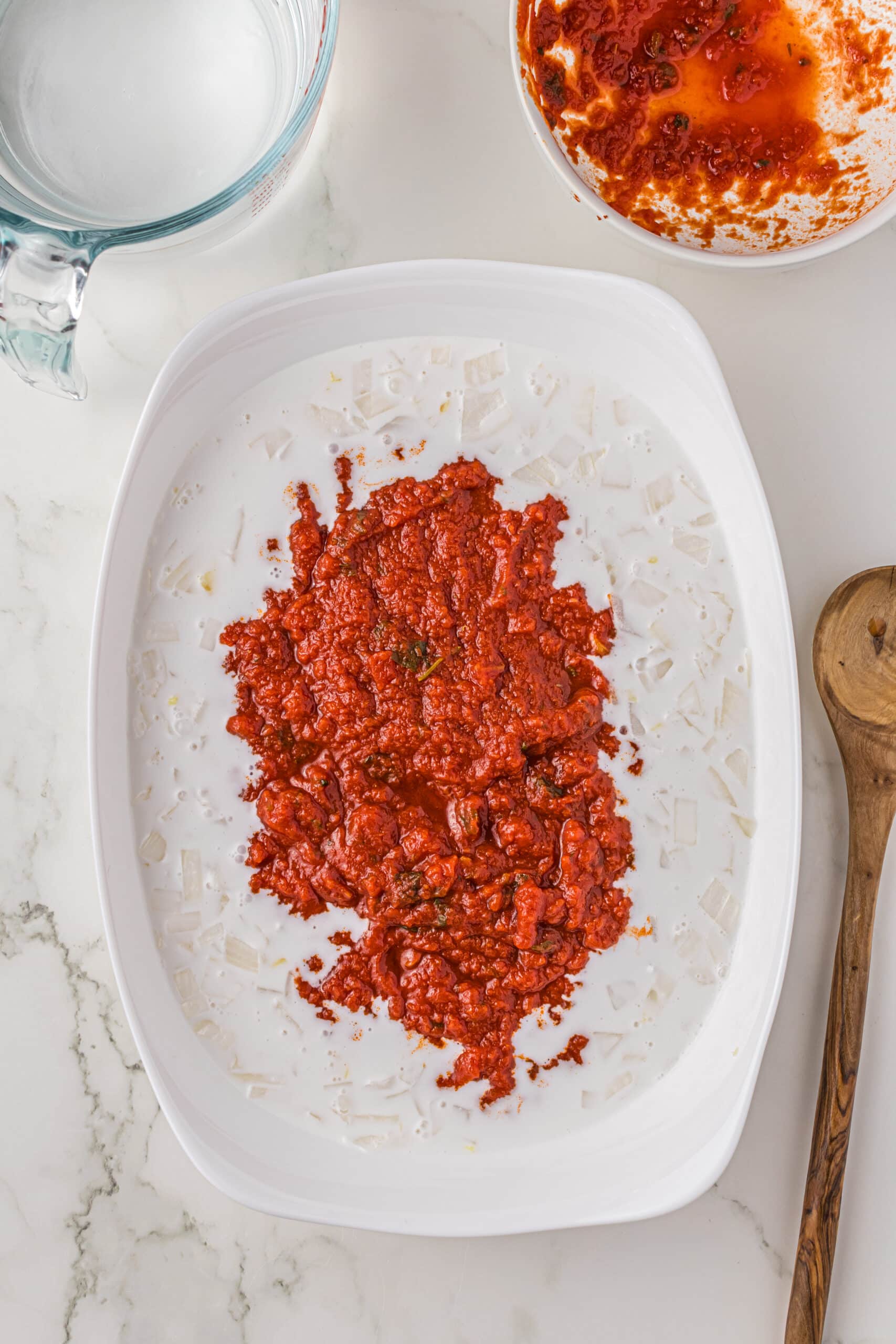 A white casserole dish filled with marinara and coconut milk. Around the dish is a glass pitcher, a white bowl that was filled with sauce, an a wooden spoon.