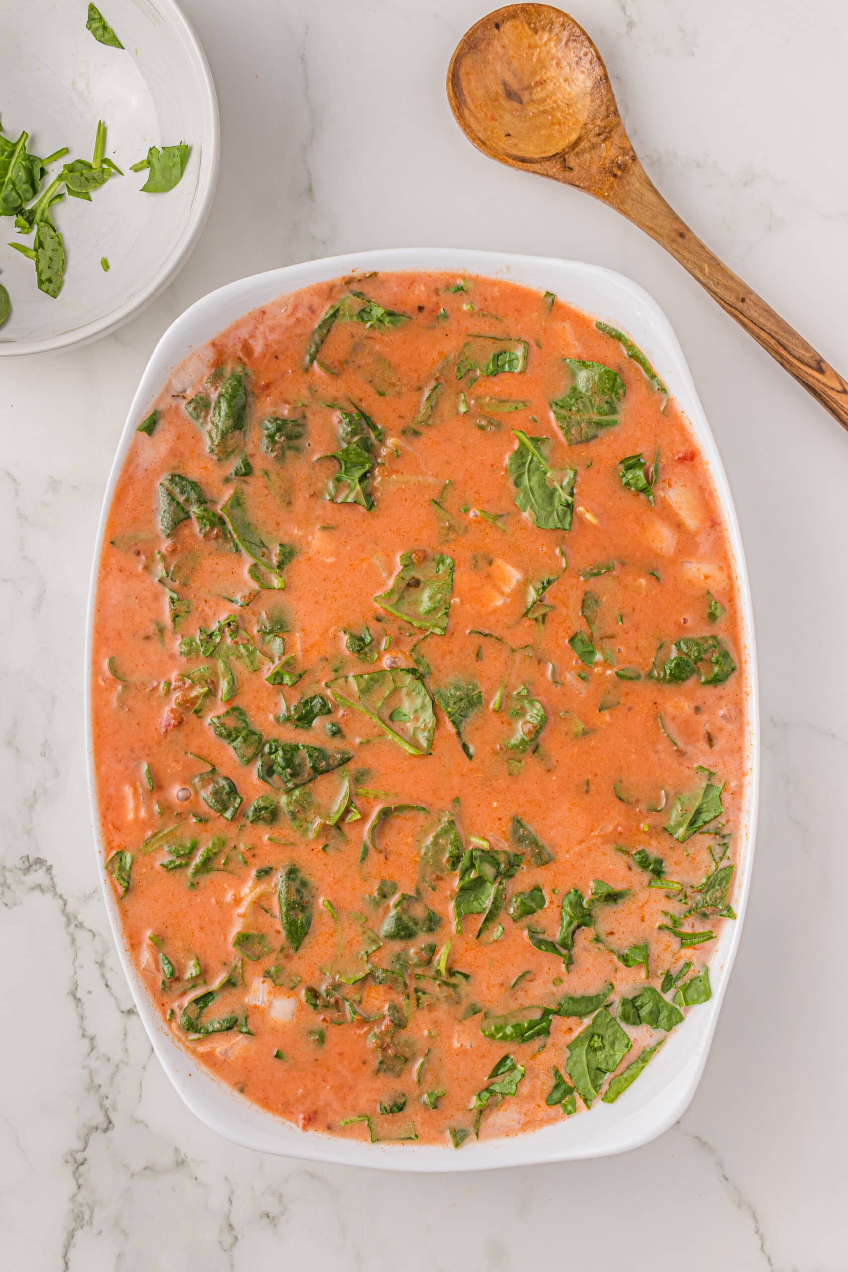 A white casserole dish filled with marinara, spinach, bow tie pasta, coconut milk, onion, and spices. Above the dish is a wooden spoon and a small white bowl with remaining fresh basil.