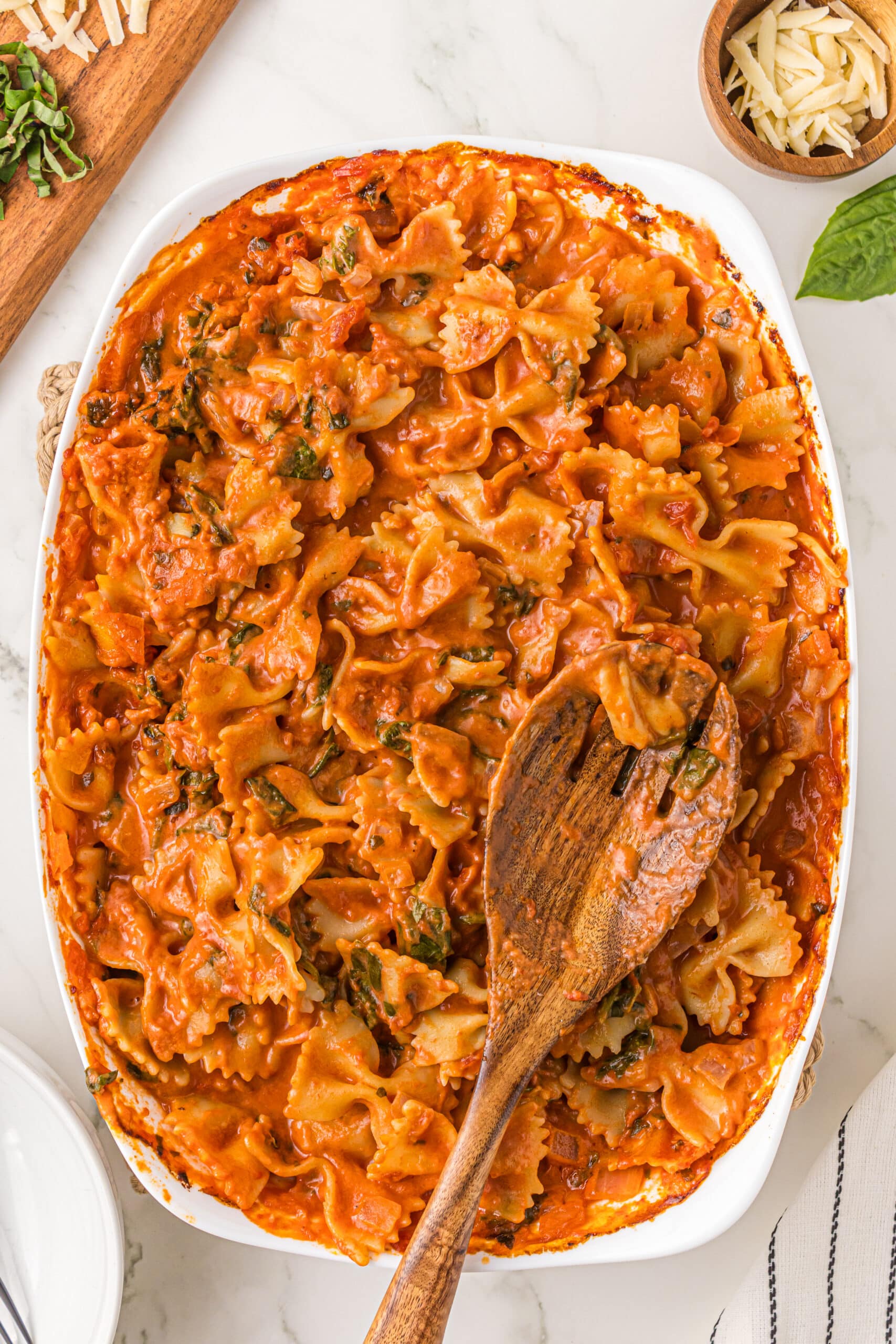 A white casserole dish filled with no boil creamy pasta bake with a wooden serving spoon resting on top of the dish. Around the bowl is a brown cutting board and a small wooden bowl of parmesan cheese with a piece of basil next to it.