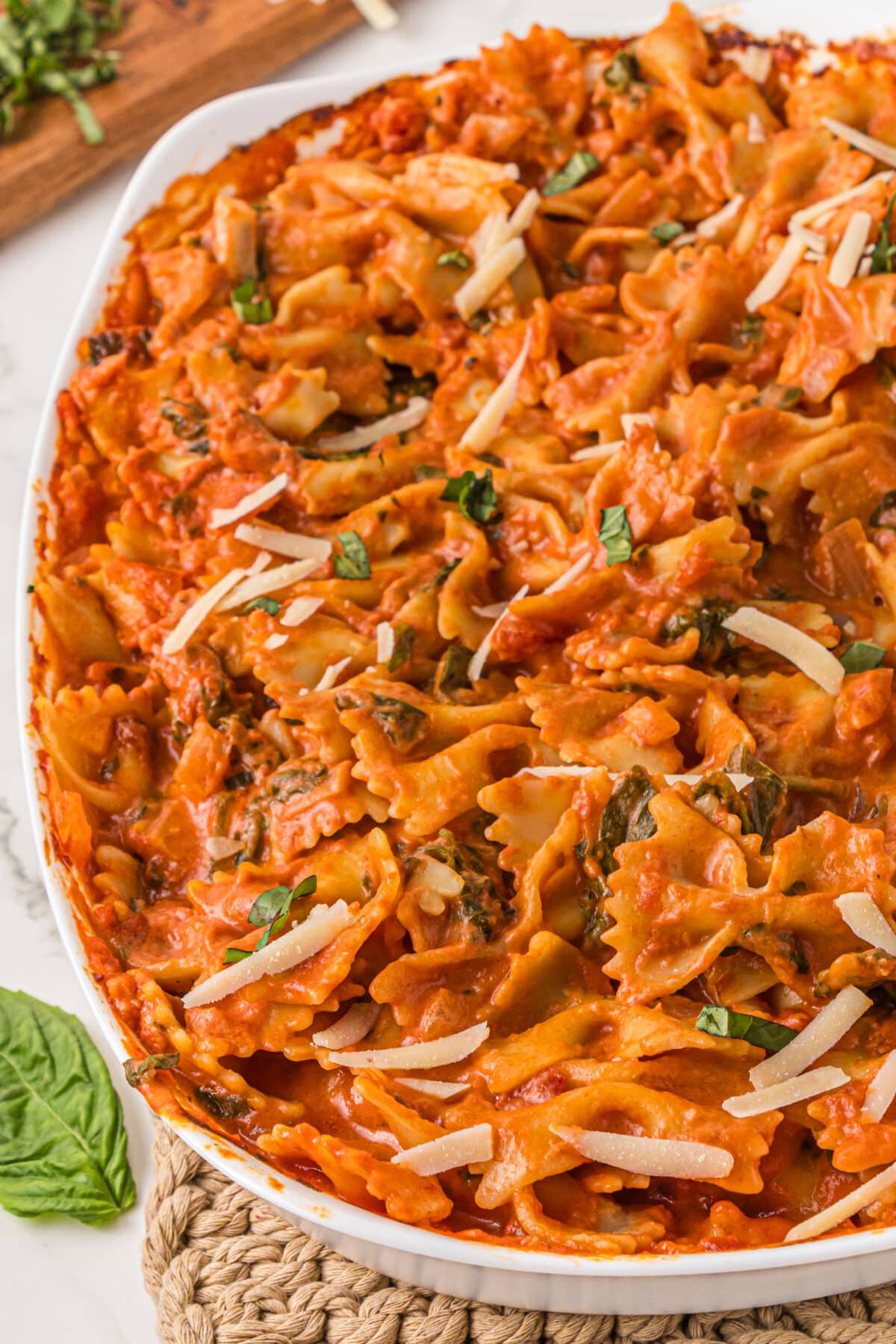 A white casserole dish rests on a placemat filled with no boil creamy pasta bake. In the background is a wooden cutting board with fresh chopped basil on it.