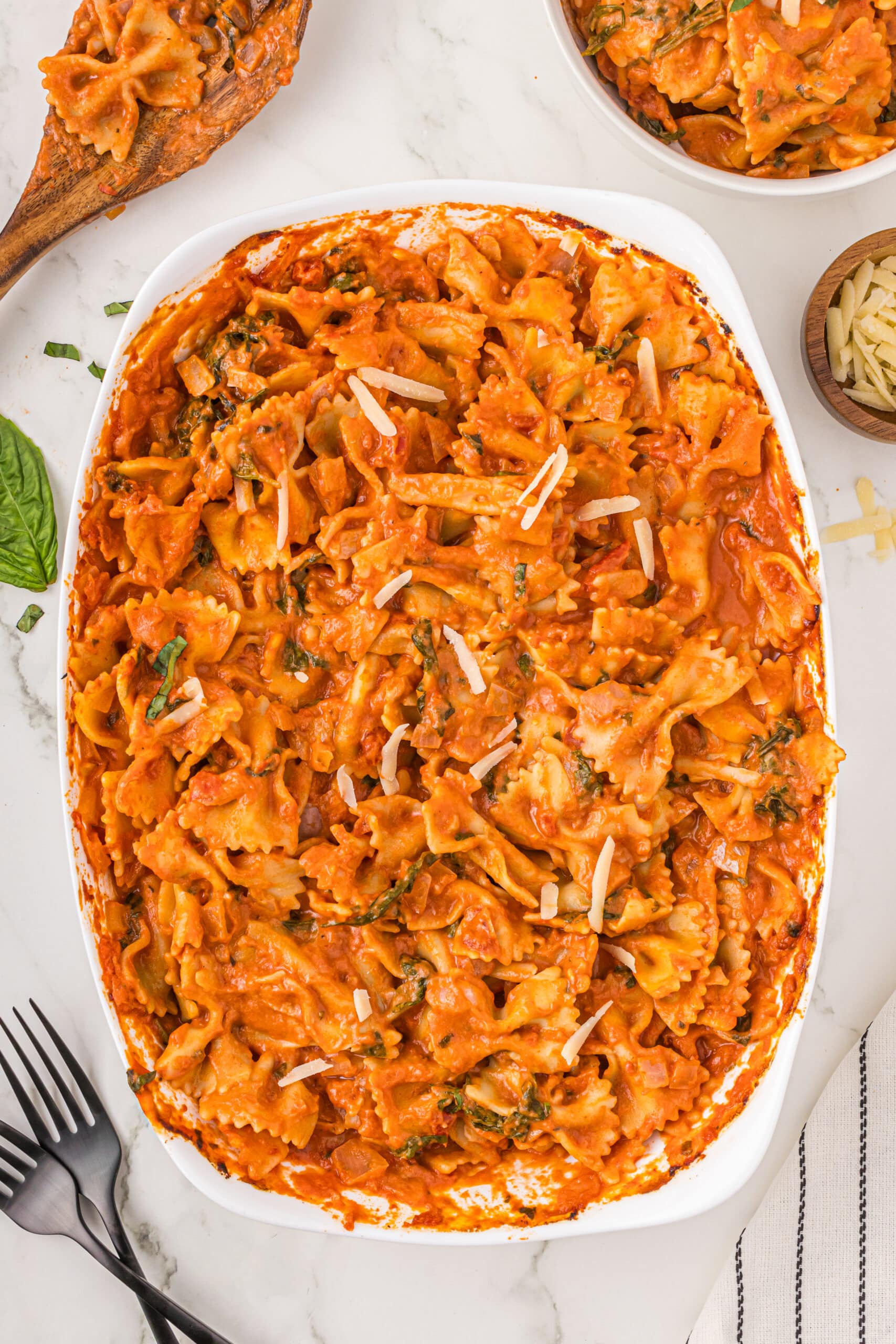 A white casserole dish filled with no-boil pasta bake garnished with parmesan shreds. Around the dish is a wooden spoon covered in sauce, a small white bowl filled with the pasta, a very small wooden bowl of parmesan cheese, two black forks, and a striped towel.