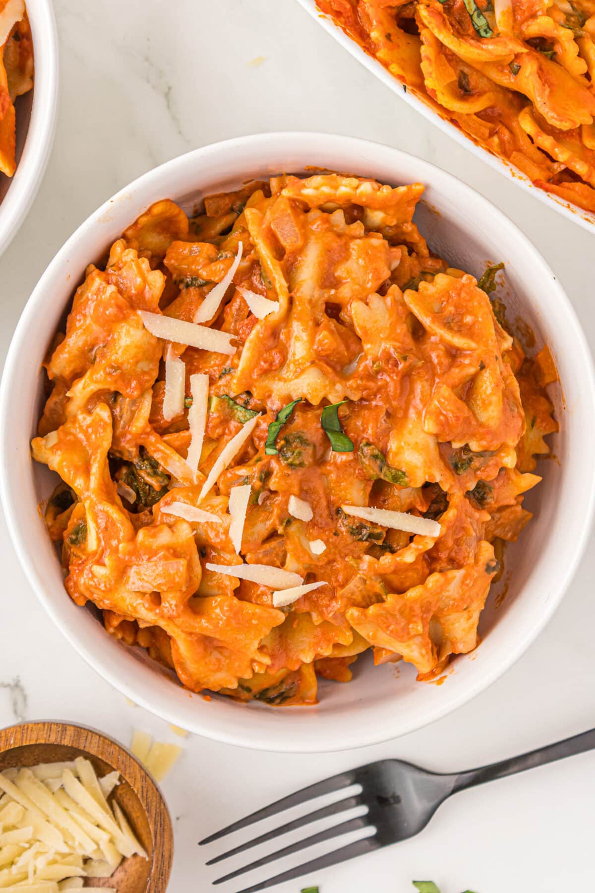 A small white bowl with a serving of no boil creamy pasta bake in it topped with basil and parmesan shreds. Around the bowl is a black fork, a small wooden bowl of cheese, and the edge of the casserole dish filled with remaining pasta.
