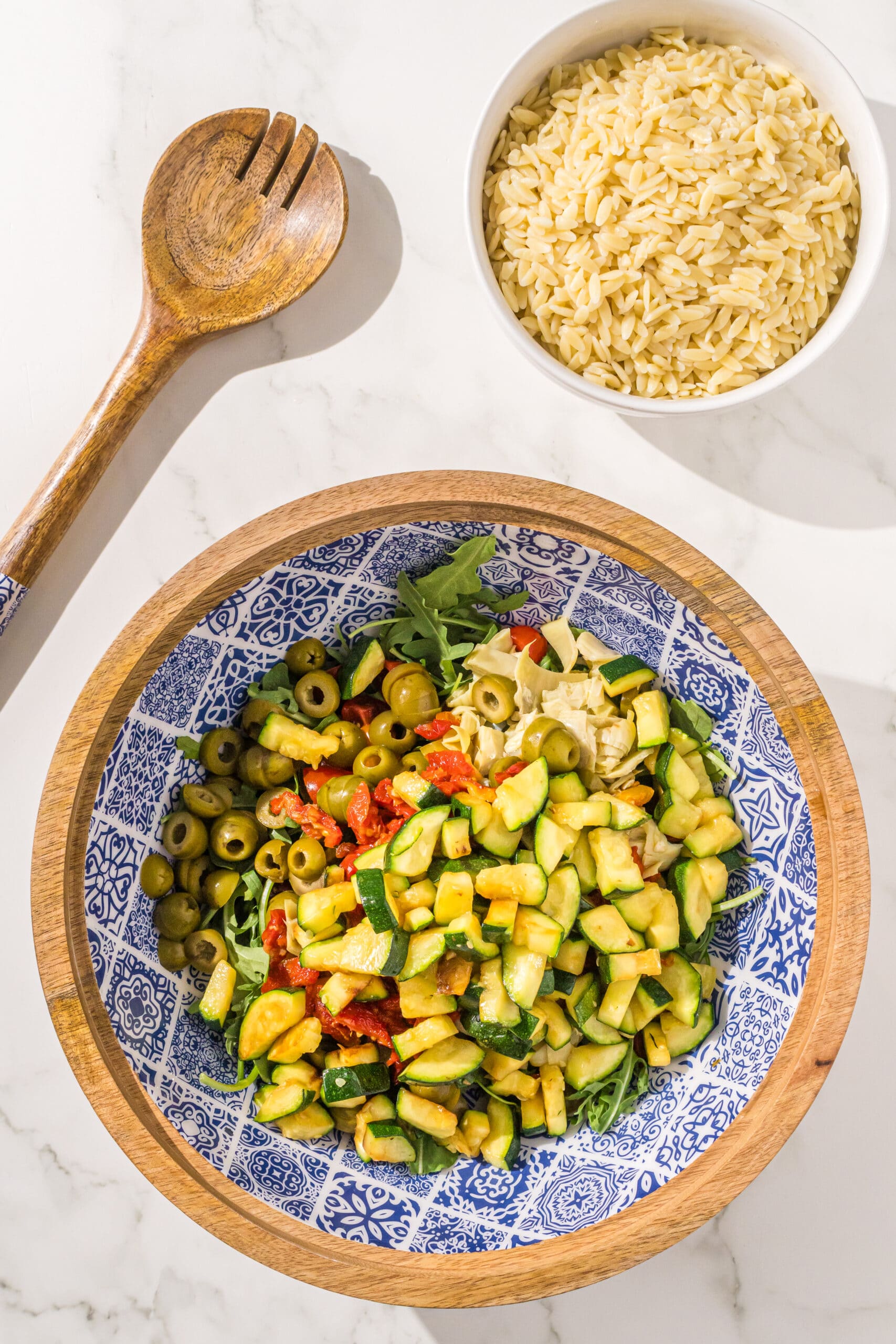 A large wooden salad bowl filled with arugula, cherry tomatoes, green olives, artichokes, sundried tomatoes, and sauteed zucchini. Beside the bowl is a wooden spoon and a medium sized bowl with cooked orzo in it.