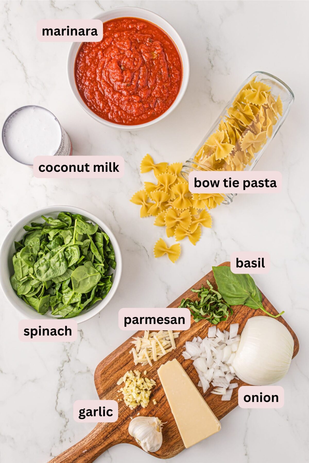 Ingredients needed to make no boil creamy pasta laid out and labeled including: marinara, coconut milk, bow tie pasta, spinach, basil, parmesan, garlic, and onion.