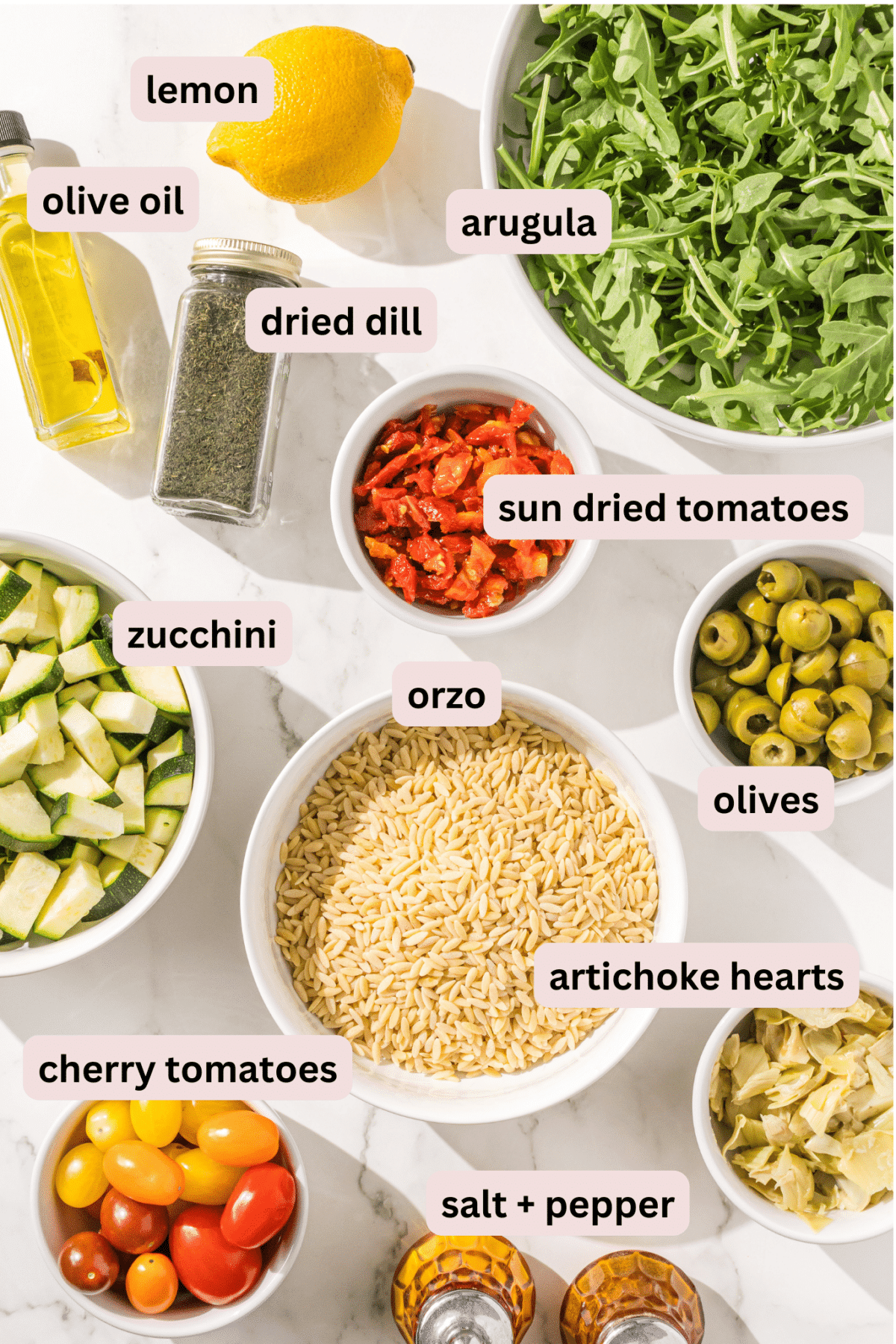 Ingredients needed to make mediterranean orzo salad laid out on the counter and labeled including: arugula, lemon, olive oil, dried dill, sundried tomatoes, olives, zucchini, orzo, artichoke hearts, cherry tomatoes, salt and pepper.