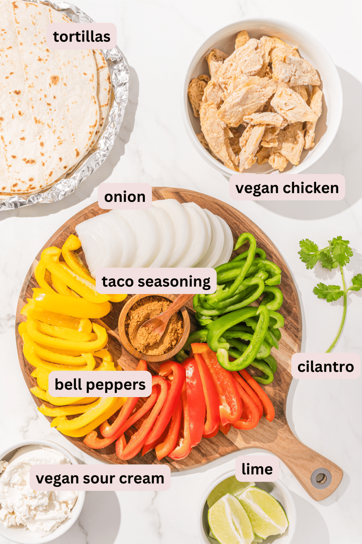 Ingredients laid out on a countertop on plates and labeled including: tortillas, vegan chicken, taco seasoning, onion, bell peppers, cilantro, vegan sour cream, and lime wedges.