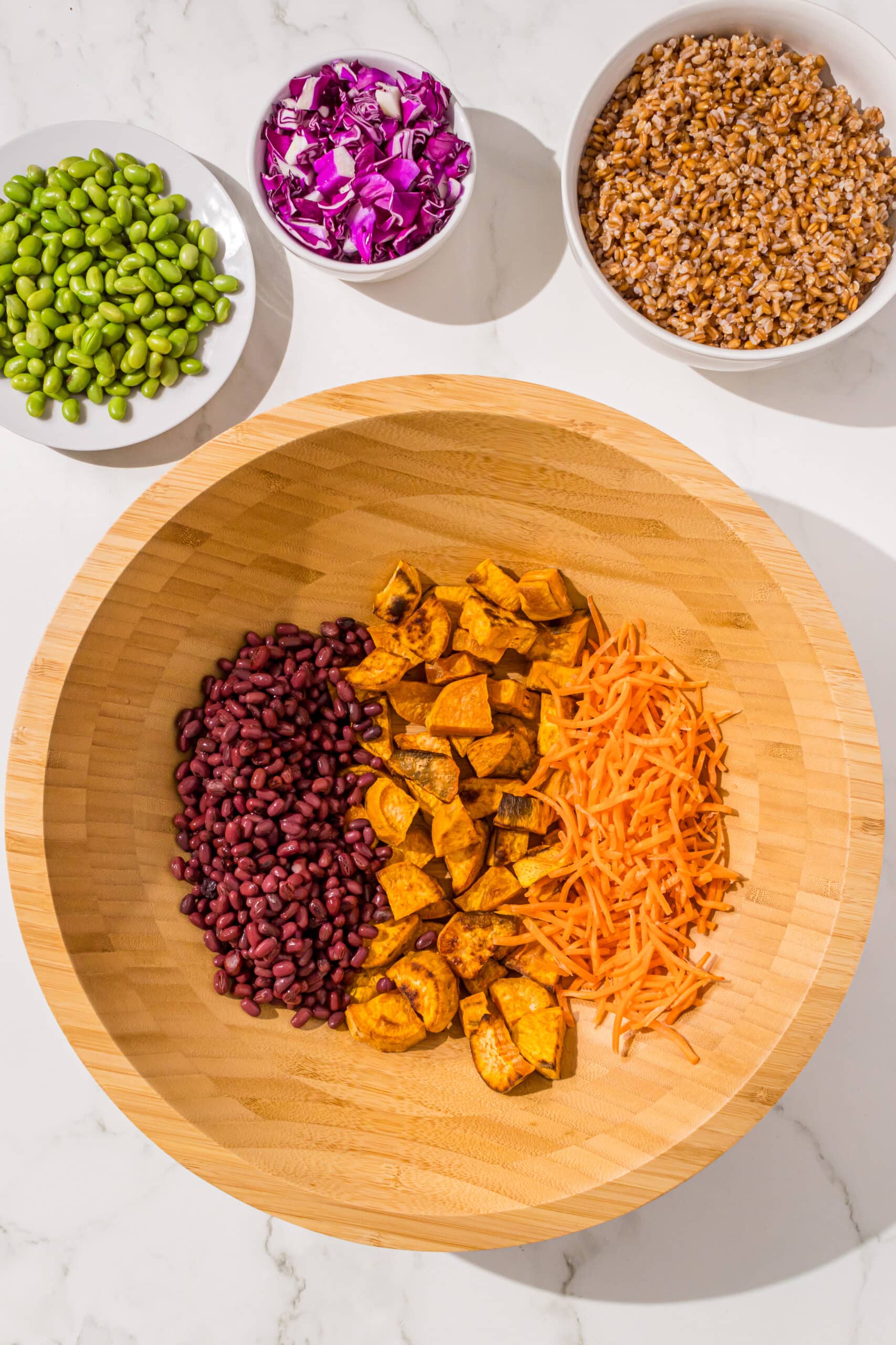 A large wooden bowl filled with adzuki beans, roasted sweet potato cubes, and shredded carrots. Beside the salad bowl are three other small bowls with edamame, purple cabbage, and farro.