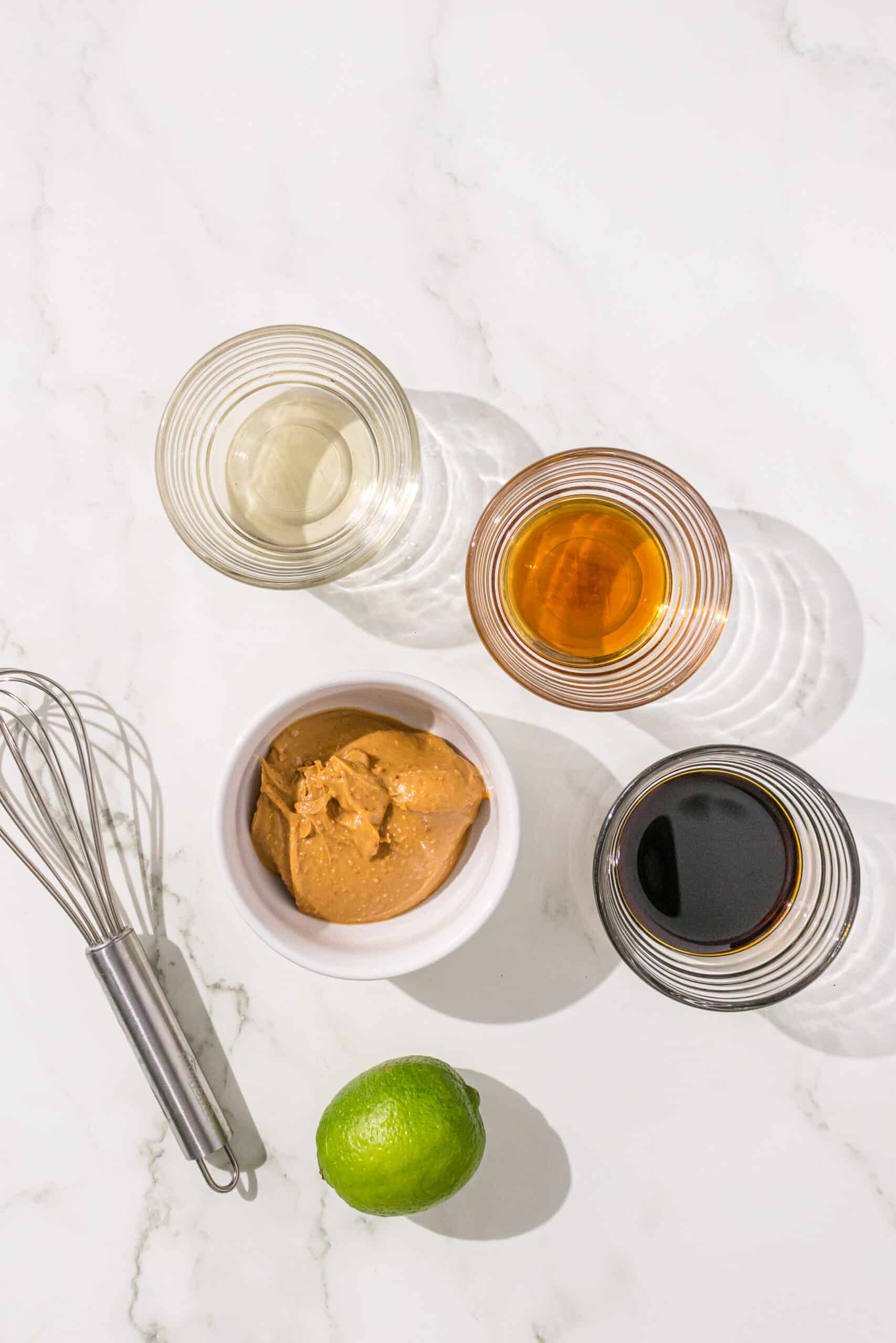 Ingredients needed to make peanut dressing in small glass bowls including: soy sauce, peanut butter, sesame oil, rice vinegar, a lime, and a small whisk.