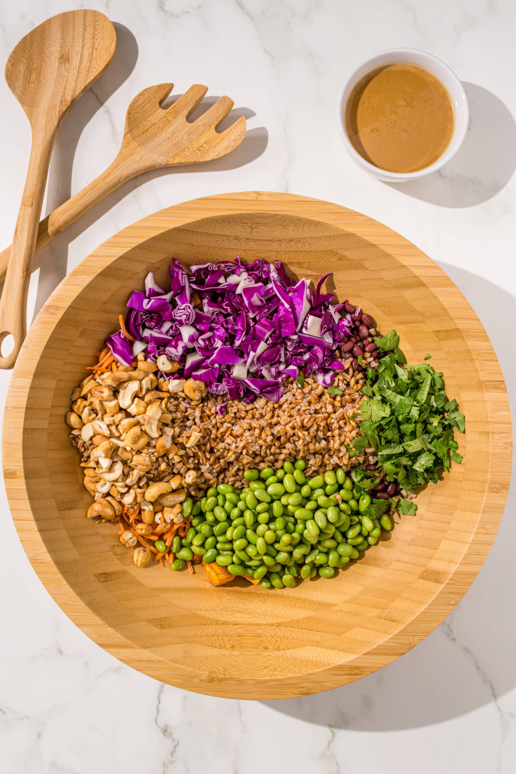 A large wooden salad bowl with farro, edamame, green onion, purple cabbage, cashews, shredded carrot, adzuki beans, and roasted sweet potatoes. Beside the bowl is a set of wooden tongs and a small bowl of peanut dressing.