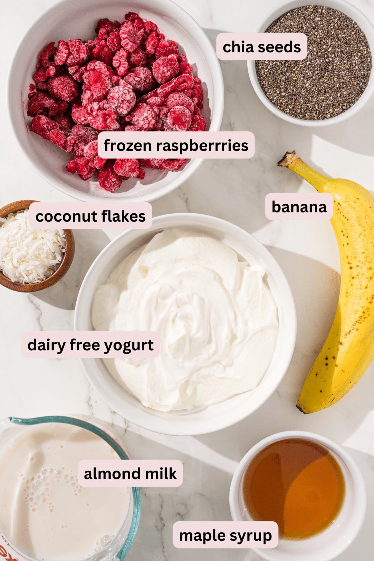 Ingredients needed to make raspberry chia pudding laid out on a marble counter and labeled including: frozen raspberries, chia seeds, coconut flakes, banana, dairy free yogurt, maple syrup, and almond milk.