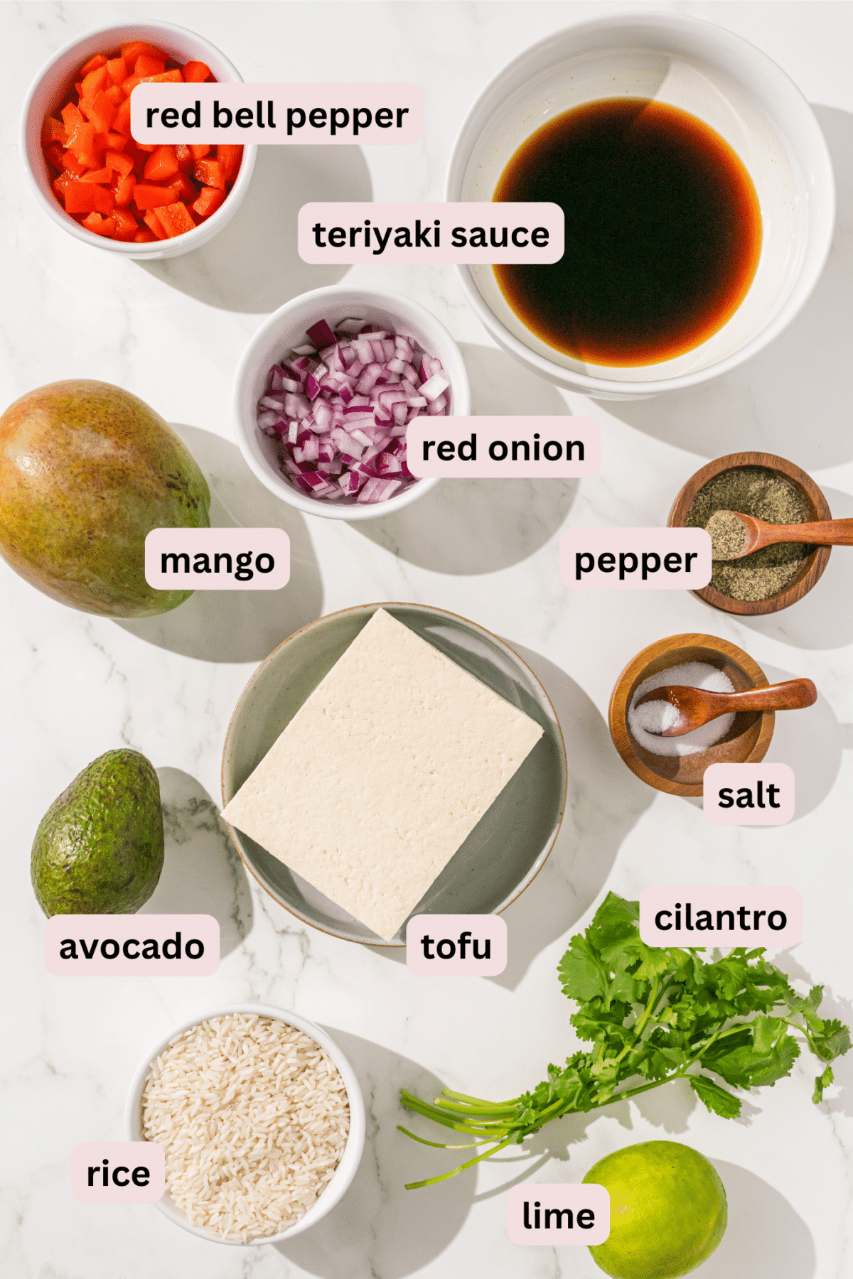 All ingredients needed for tofu dinner bowls laid out on a marble counter and labeled including: bell pepper, teriyaki sauce, red onion, mango, pepper, salt, avocado, tofu, cilantro, rice, and lime.