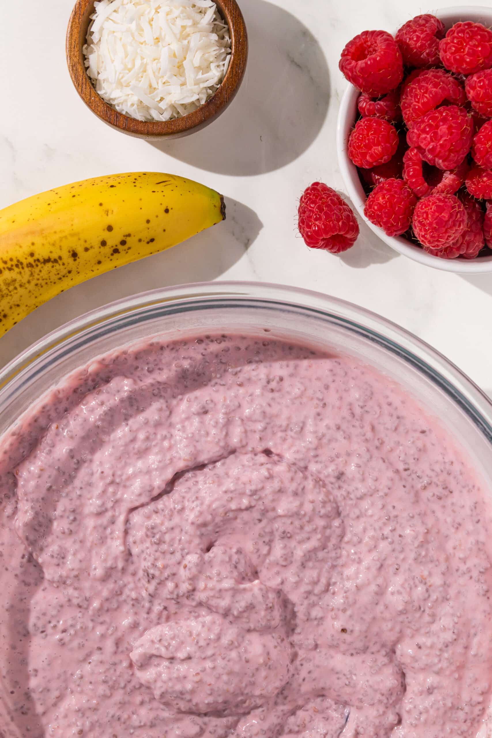 A large glass bowl of raspberry chia seed pudding with a banana, handful of raspberries, and small wooden bowl of shredded coconut above it.
