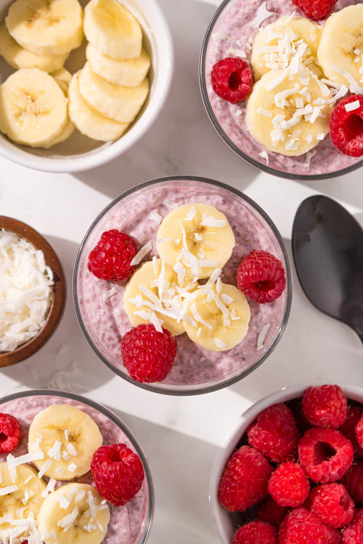 Three glass jars of raspberry chia pudding topped with raspberries, banana and coconut. To the left of the jars is a white bowl of bananas and a small wooden bowl of shredded coconut. To the right of the jars is a white bowl of raspberries and a black spoon.