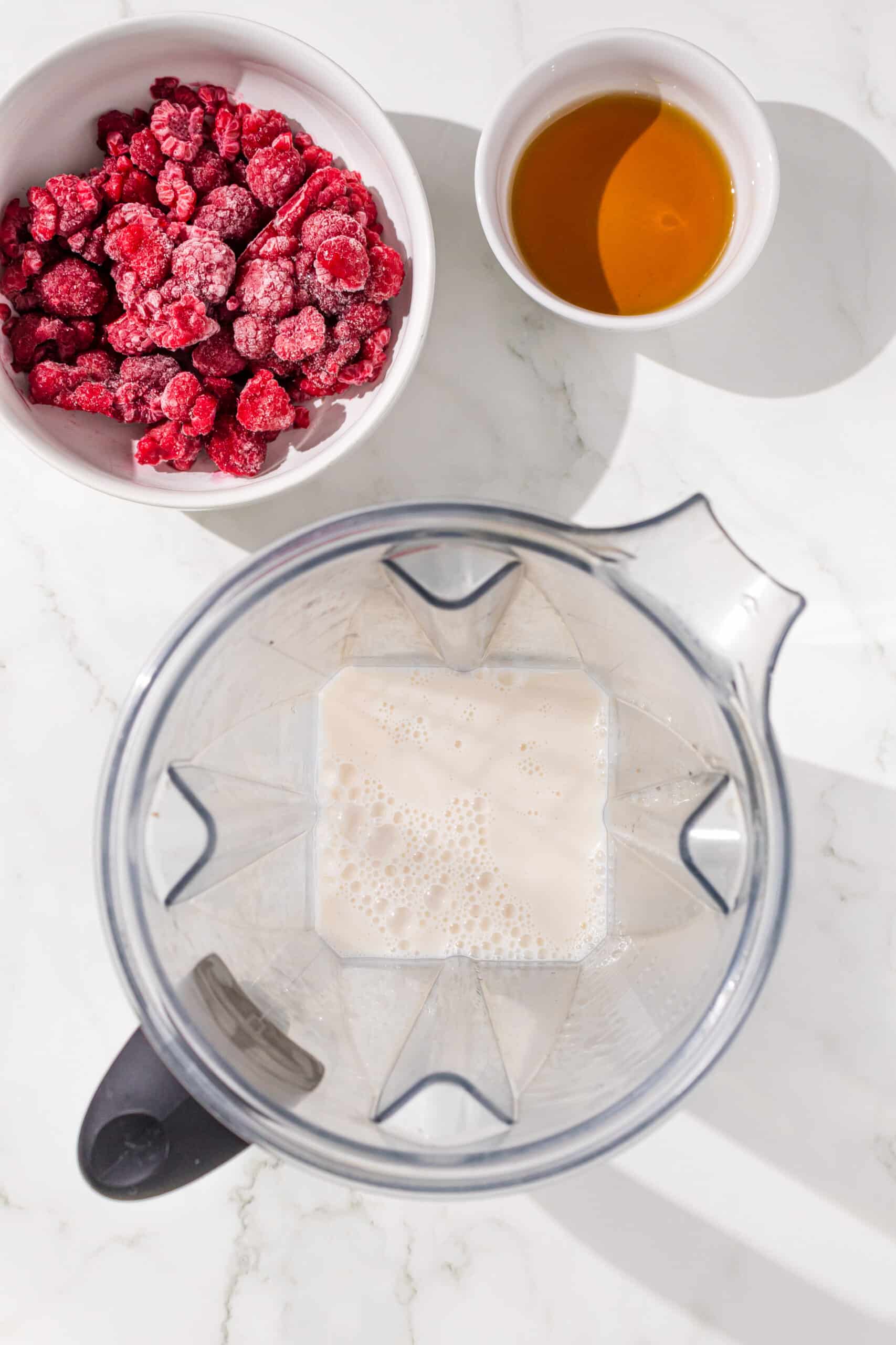 A blender with almond milk in it. Above the blender is two white bowls of frozen raspberries and maple syrup.