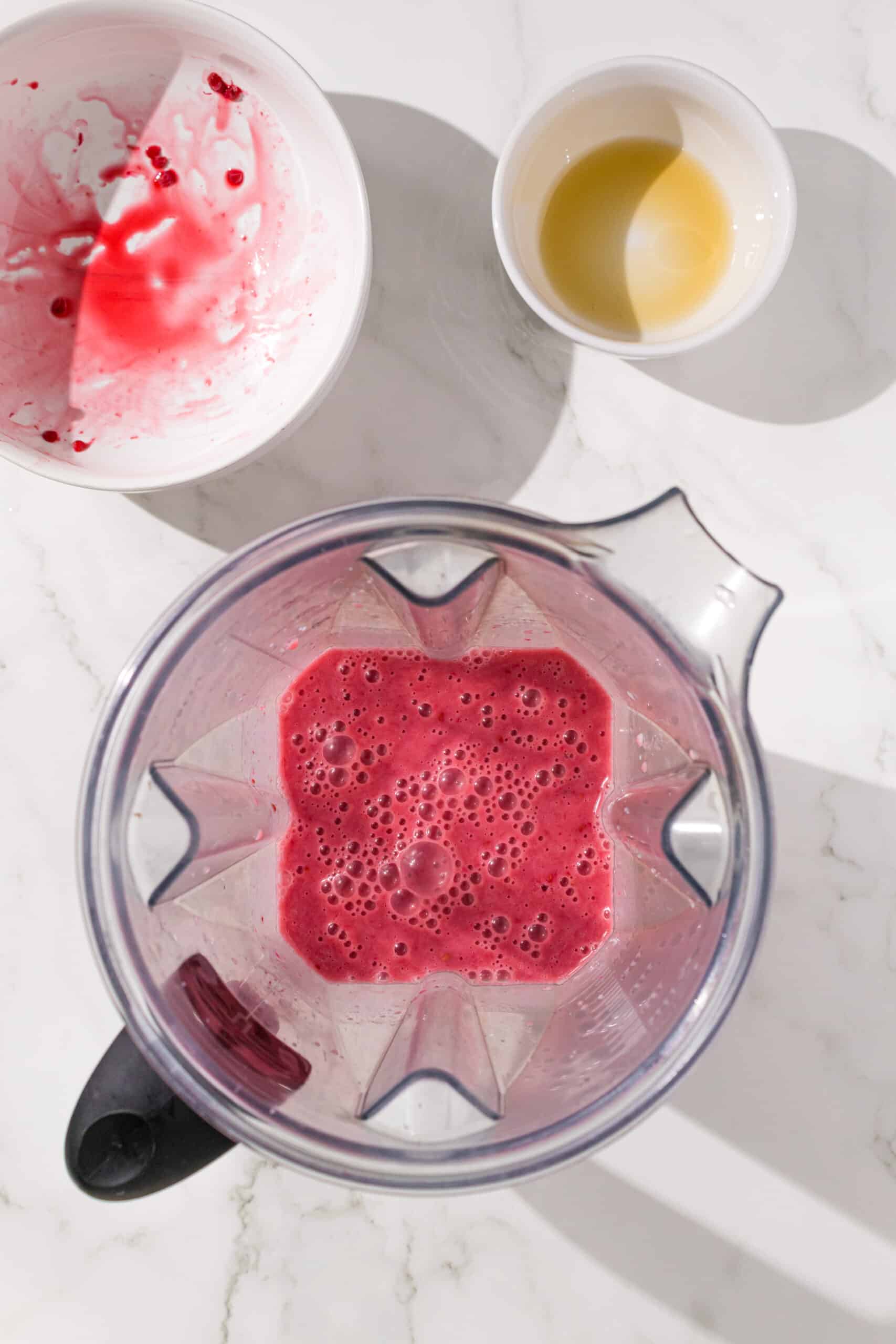 A blender filled with raspberry milk and two empty white bowls above it (that used to have raspberries and maple syrup in them).