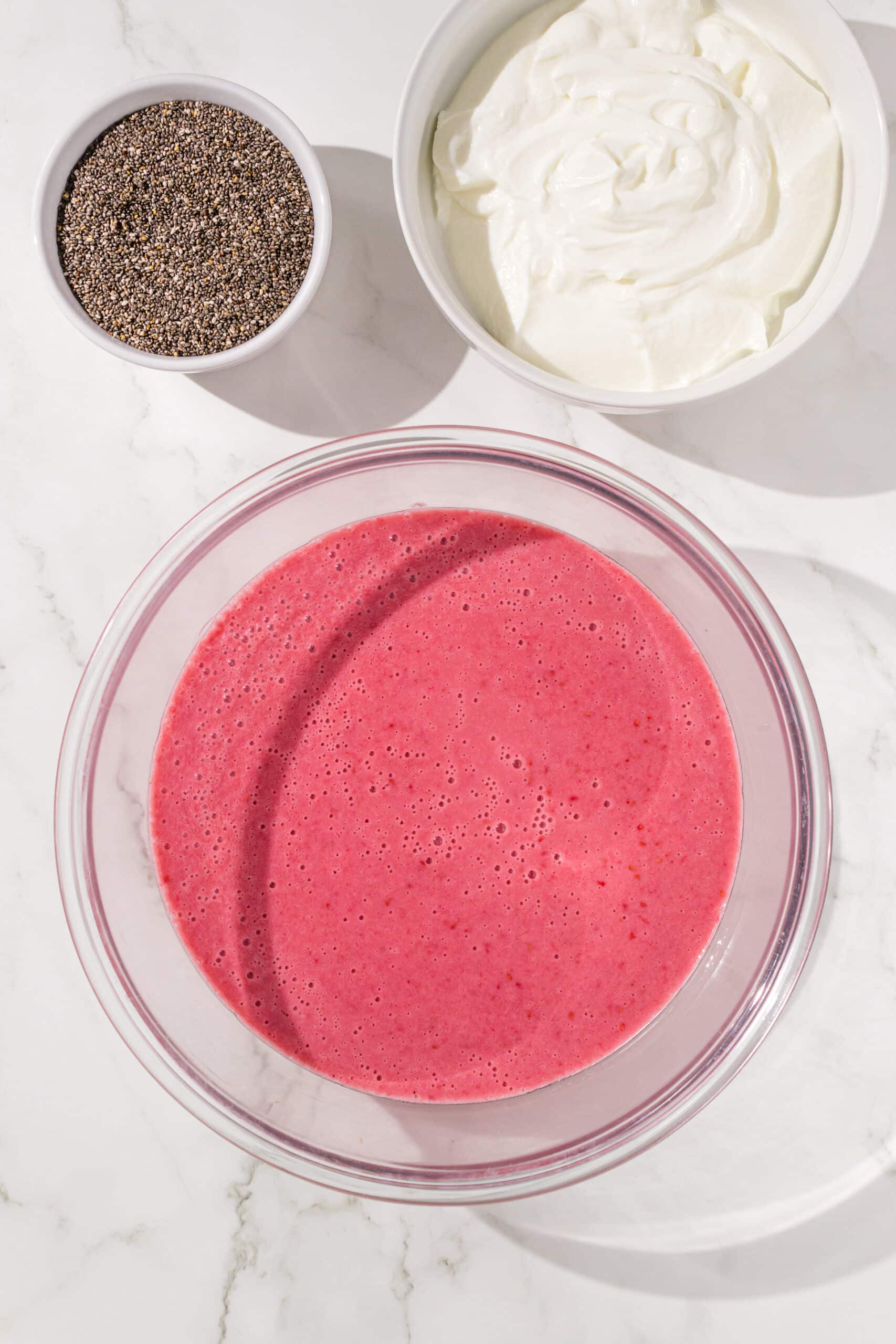 A glass bowl of raspberry milk, a white small bowl of chia seeds, and a large white bowl of dairy free yogurt.