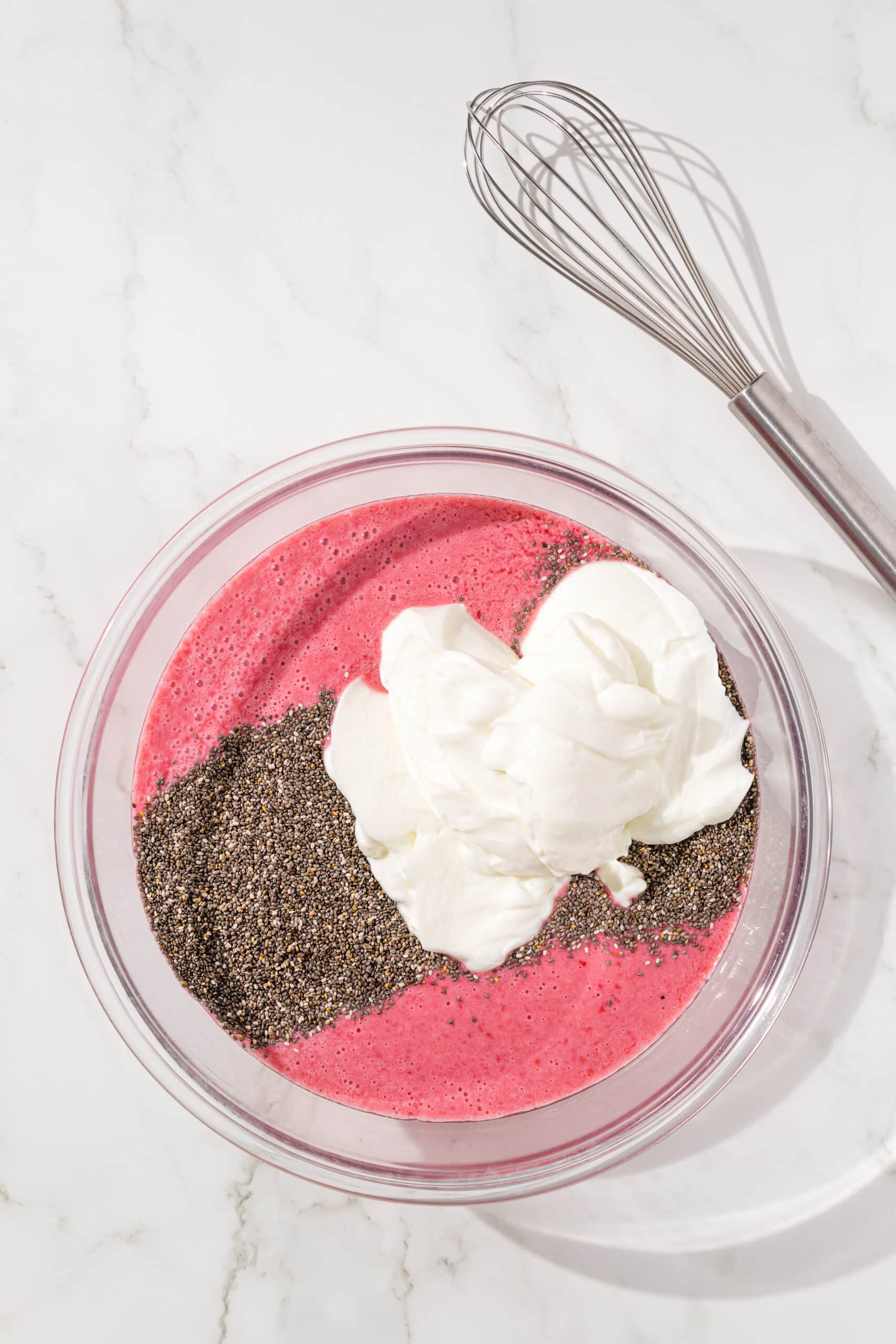 A large glass bowl of raspberry milk, chia seeds, and dairy free yogurt. There is a whisk to the right of the bowl.