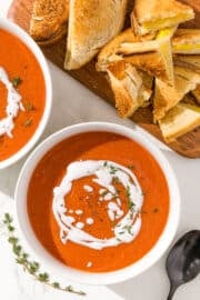 Two white bowls with red soup in them and a white sauce swirled in the center. Beside the bowls is a black metal spoon, a sprig of fresh thyme, and a cutting board with grilled cheese triangles on it.
