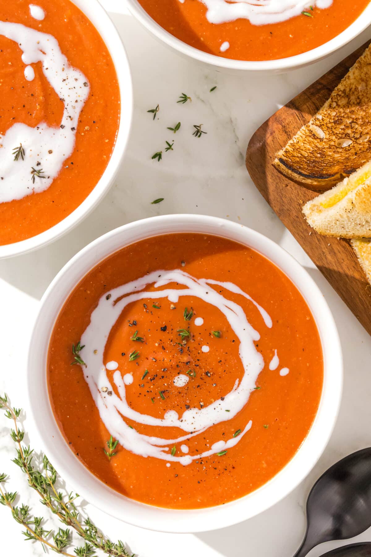 Three bowls of tomato soup garnished with a swirl of coconut cream and thyme. Beside the bowls is a set of two black metal spoons and a wood cutting board with grilled cheese triangles on it.