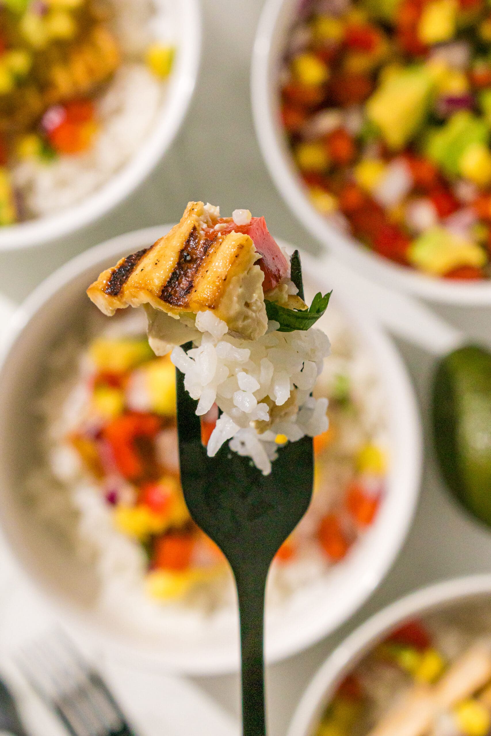 Three tofu dinner bowls in the background. In the foreground is a black fork with a bite of rice, tofu, and mango salsa on it.