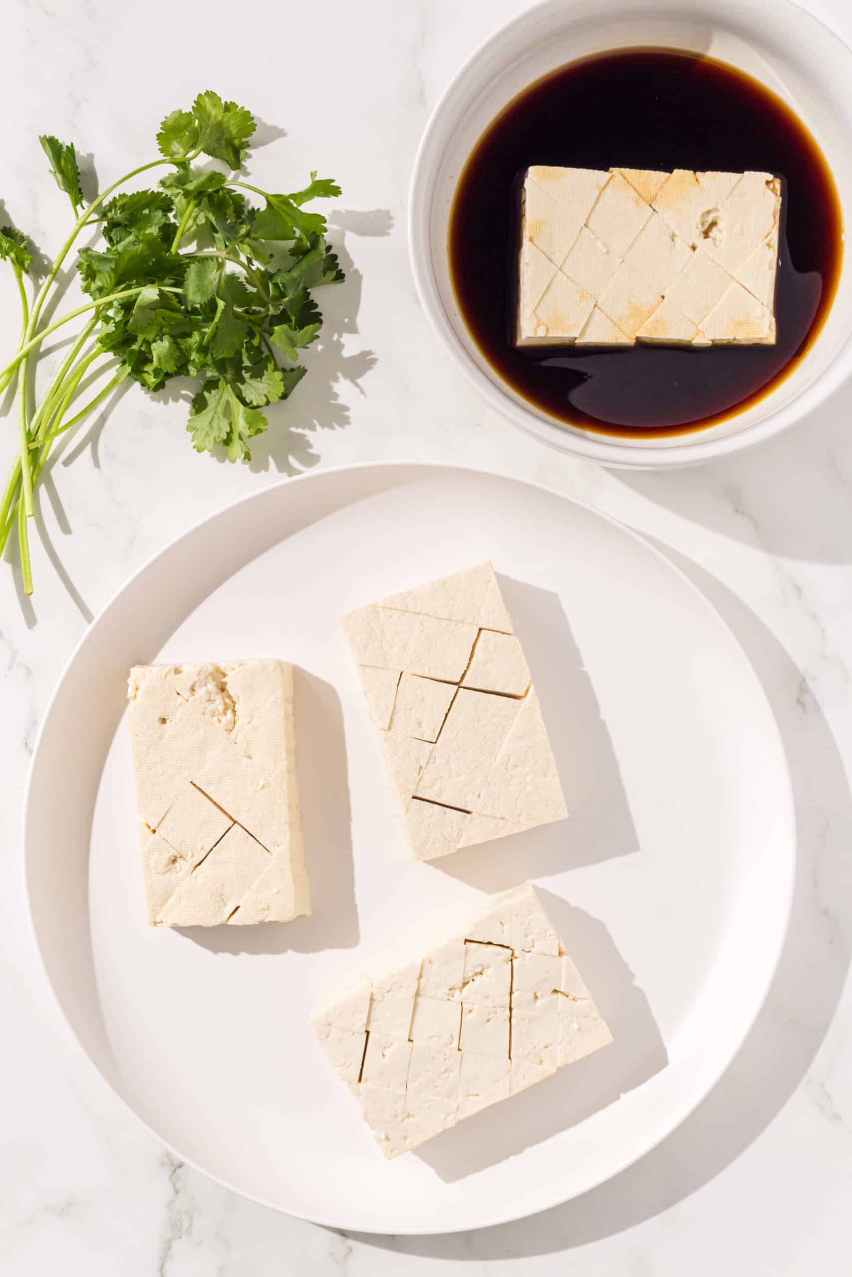3 slices of tofu on a white plate with a white bowl beside it of teriyaki sauce and 1 piece of tofu marinating in the bowl. A sprig of cilantro is to the left.
