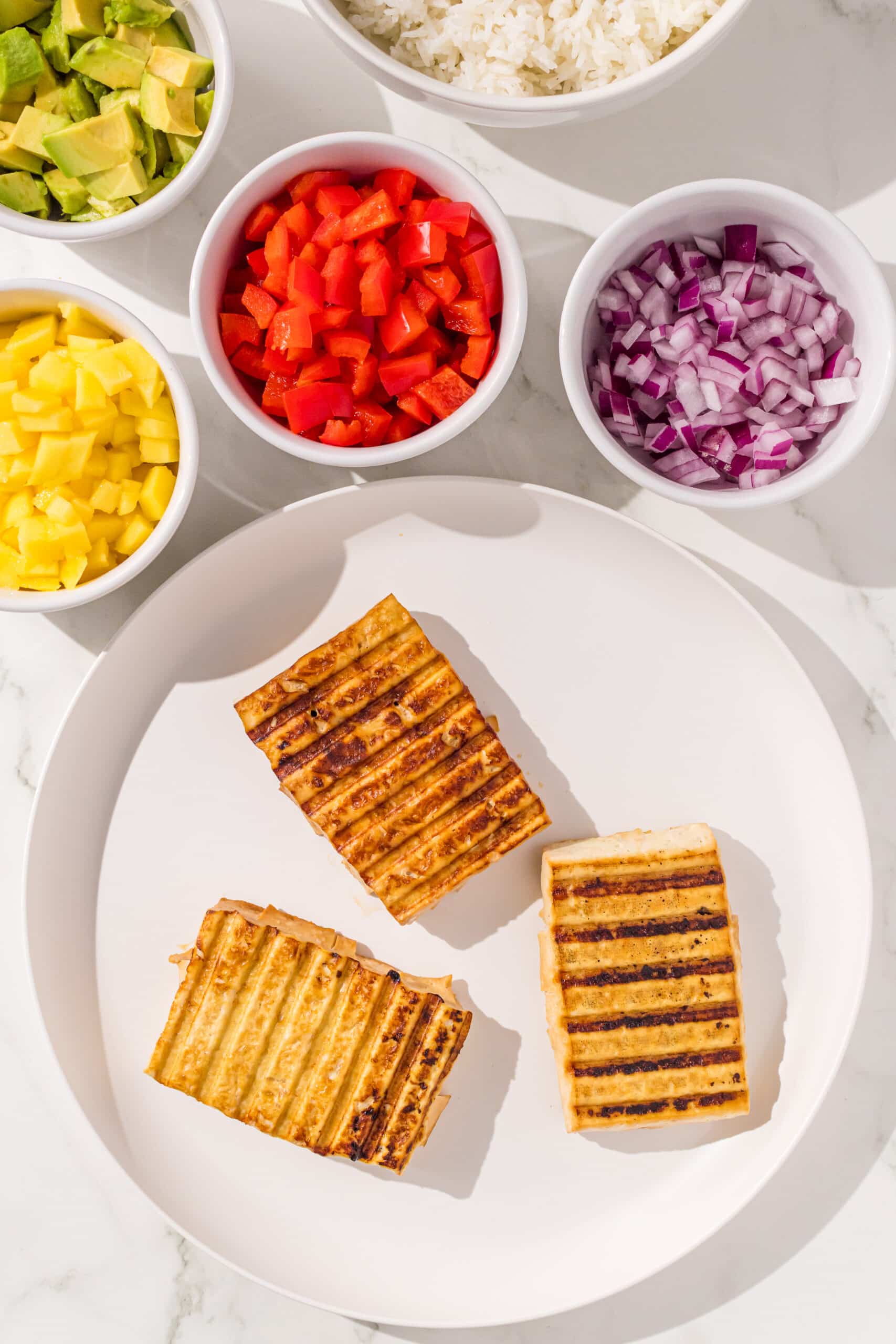 A white plate with three pieces of grilled tofu and in the back is bowls of bell pepper, onion, avocado, mango, and rice.