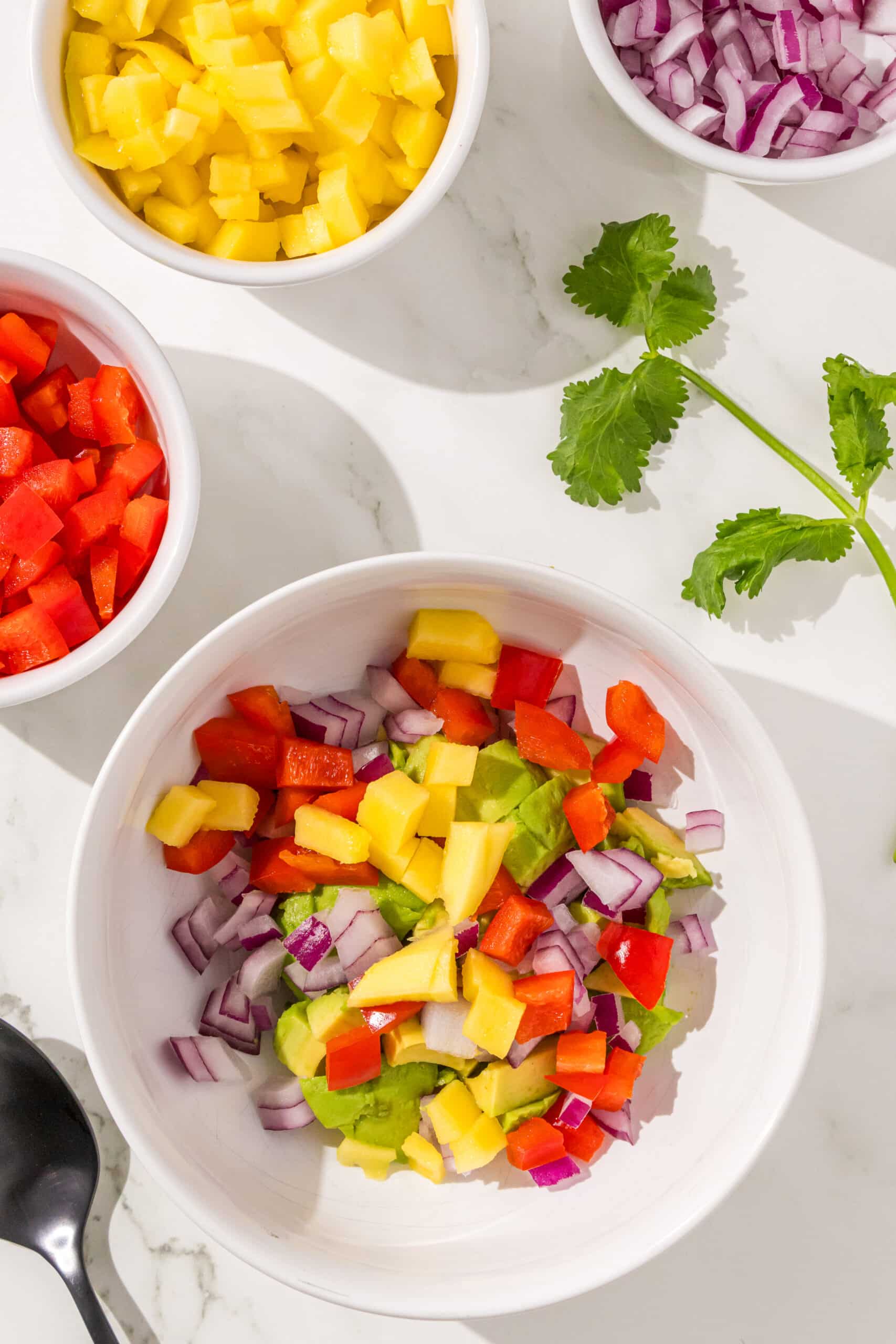 A bowl of mango salsa next to three other bowls of bell pepper, mango, and red onion. There is also a black spoon and sprig of cilantro.