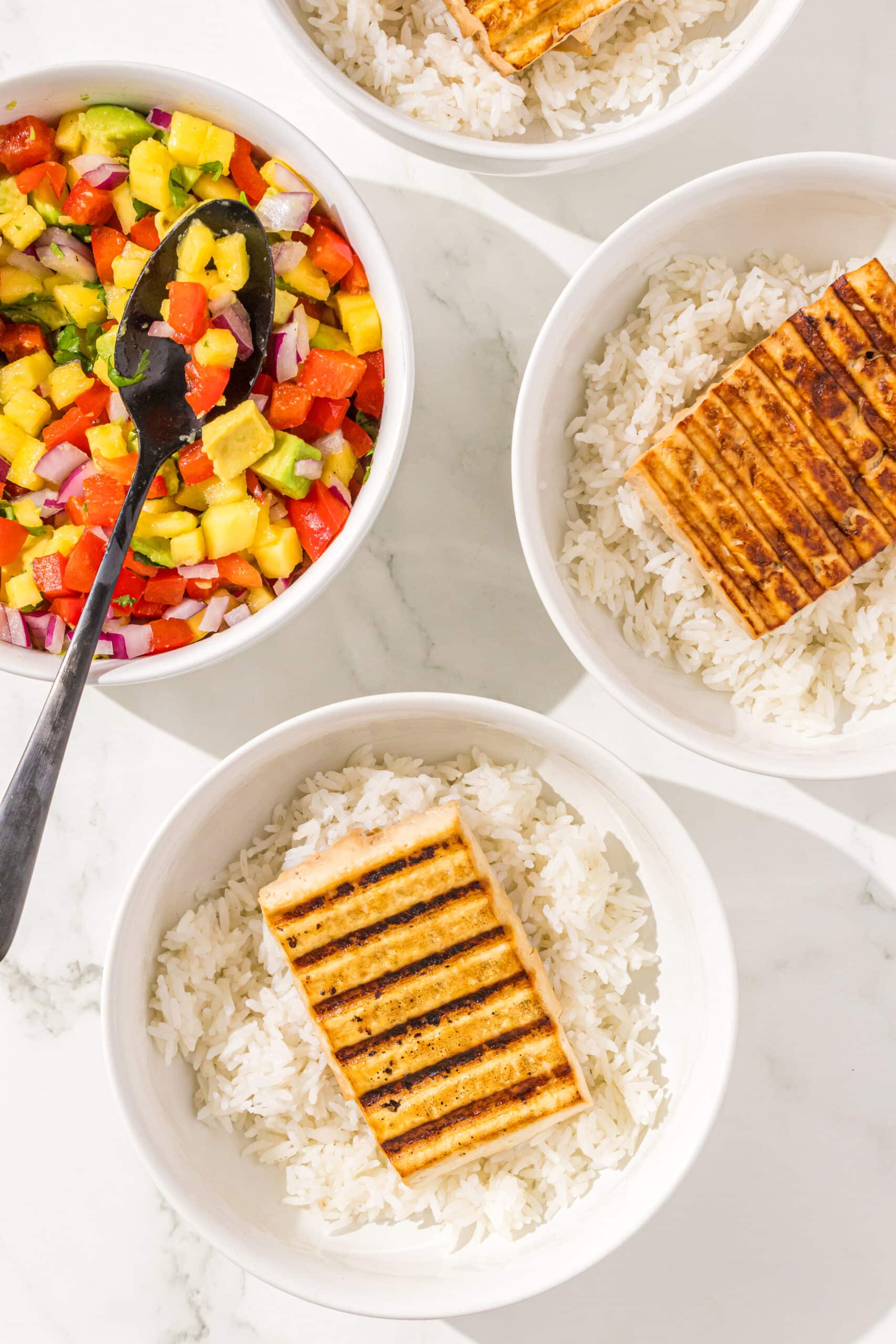 Three bowls of rice and 1 piece of tofu and one bowl of mango salsa with a black spoon in it.