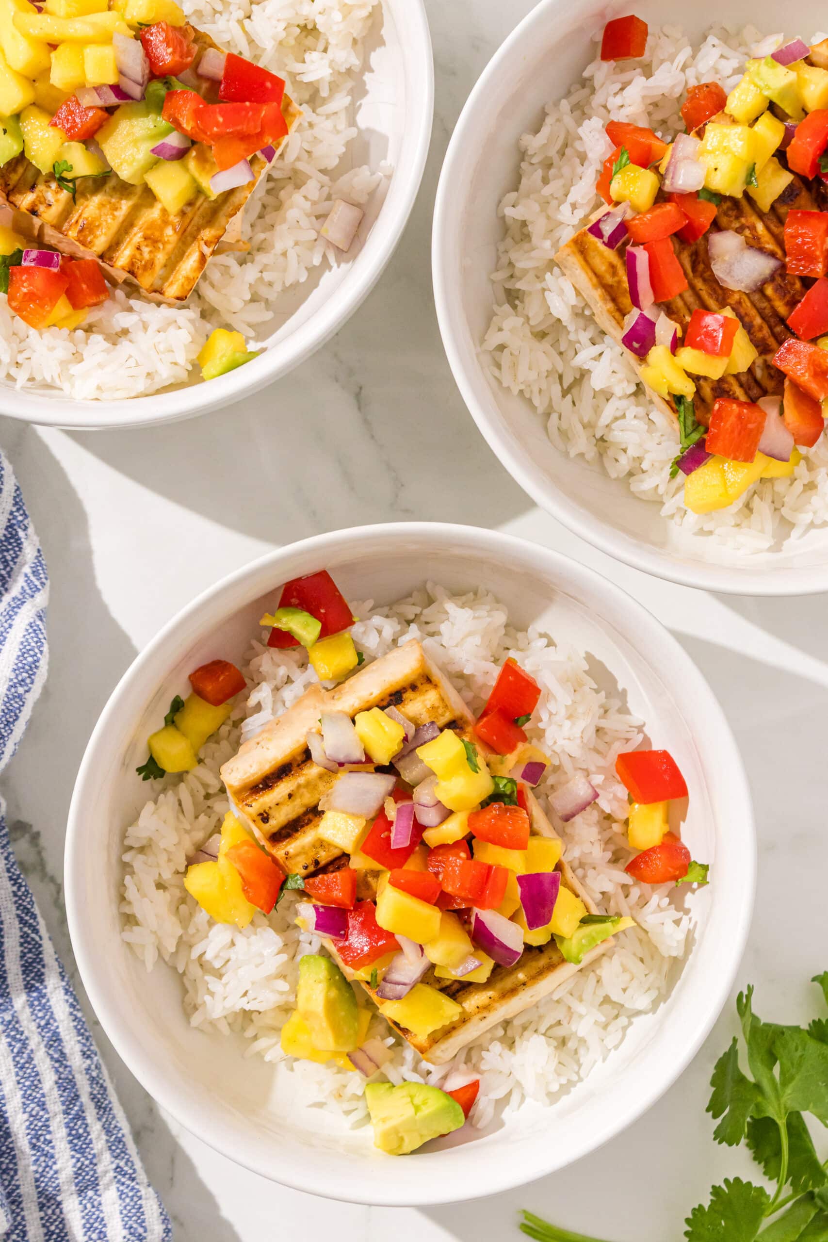 Three Tofu Dinner Bowls with a blue and white stripe towel in the corner and a sprig of cilantro.
