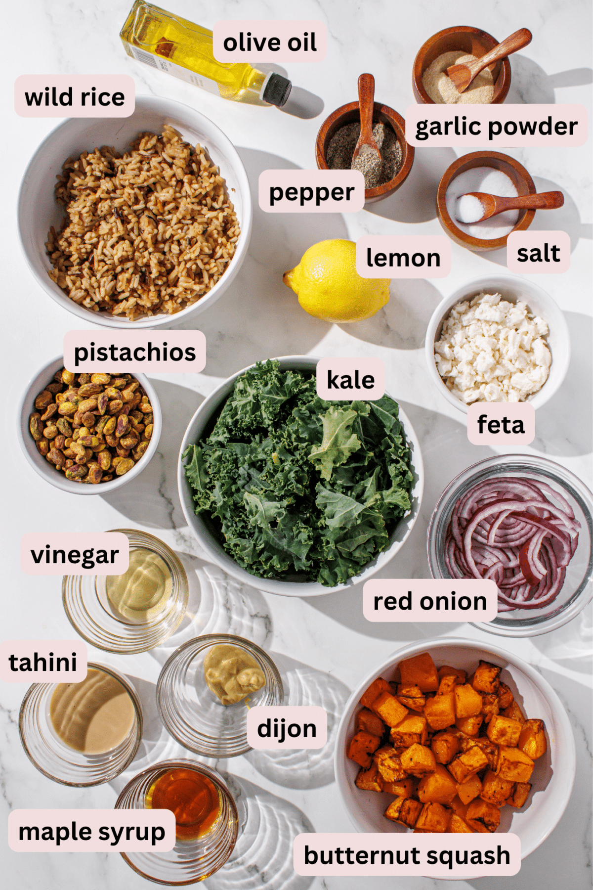 Ingredients needed to make winter wild rice salad laid out on the counter and labeled including: olive oil, garlic powder, pepper, salt, lemon, wild rice, kale, pistachios, feta, vinegar, red onion, butternut squash, dijon mustard, tahini, and maple syrup.